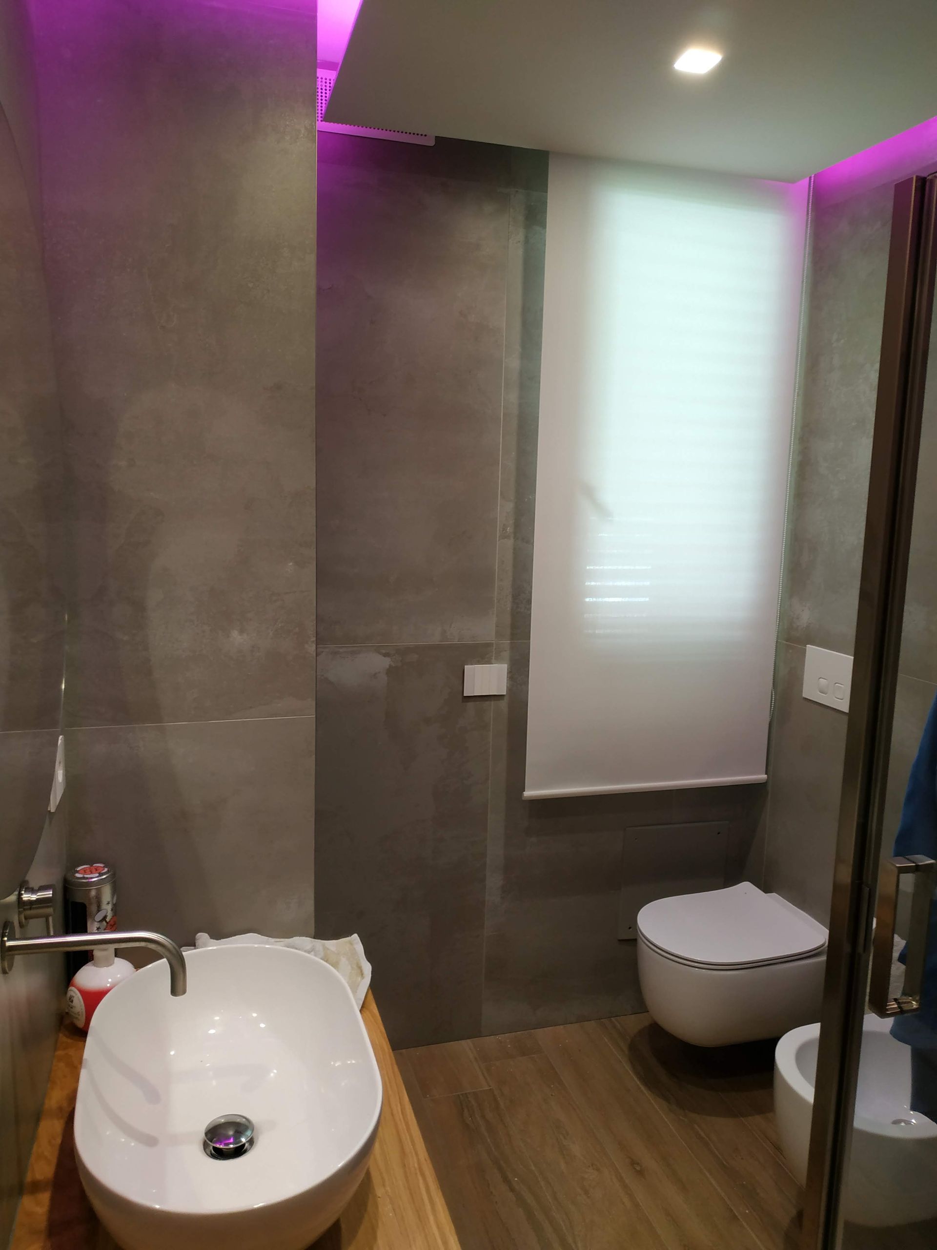Bagno moderno con piastrelle grigie, lavabo e WC bianchi, tende alle finestre e illuminazione a LED viola.