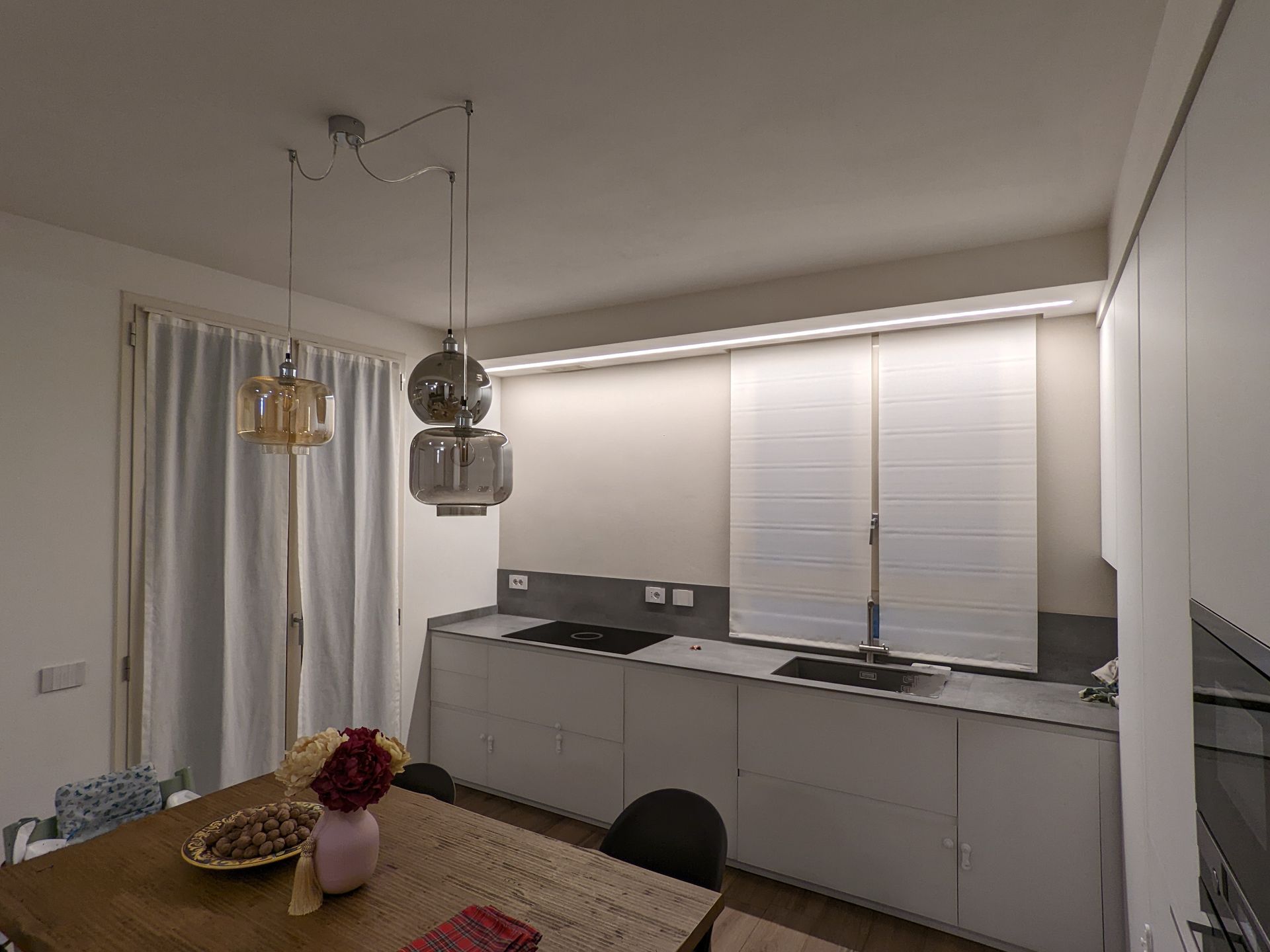 Cucina moderna con mobili bianchi, tende alle finestre, lampade a sospensione e tavolo da pranzo in legno.