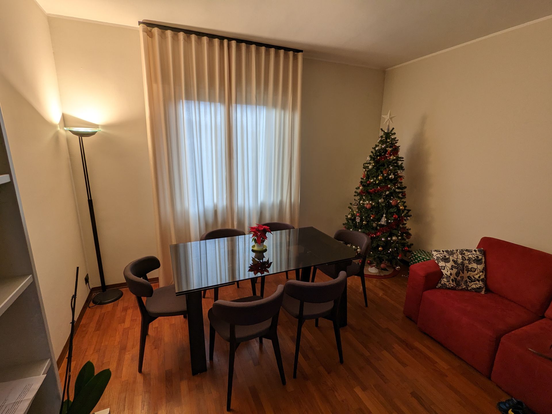 Accogliente soggiorno con tavolo da pranzo, divano rosso, lampada da terra e albero di Natale illuminato vicino a una finestra.