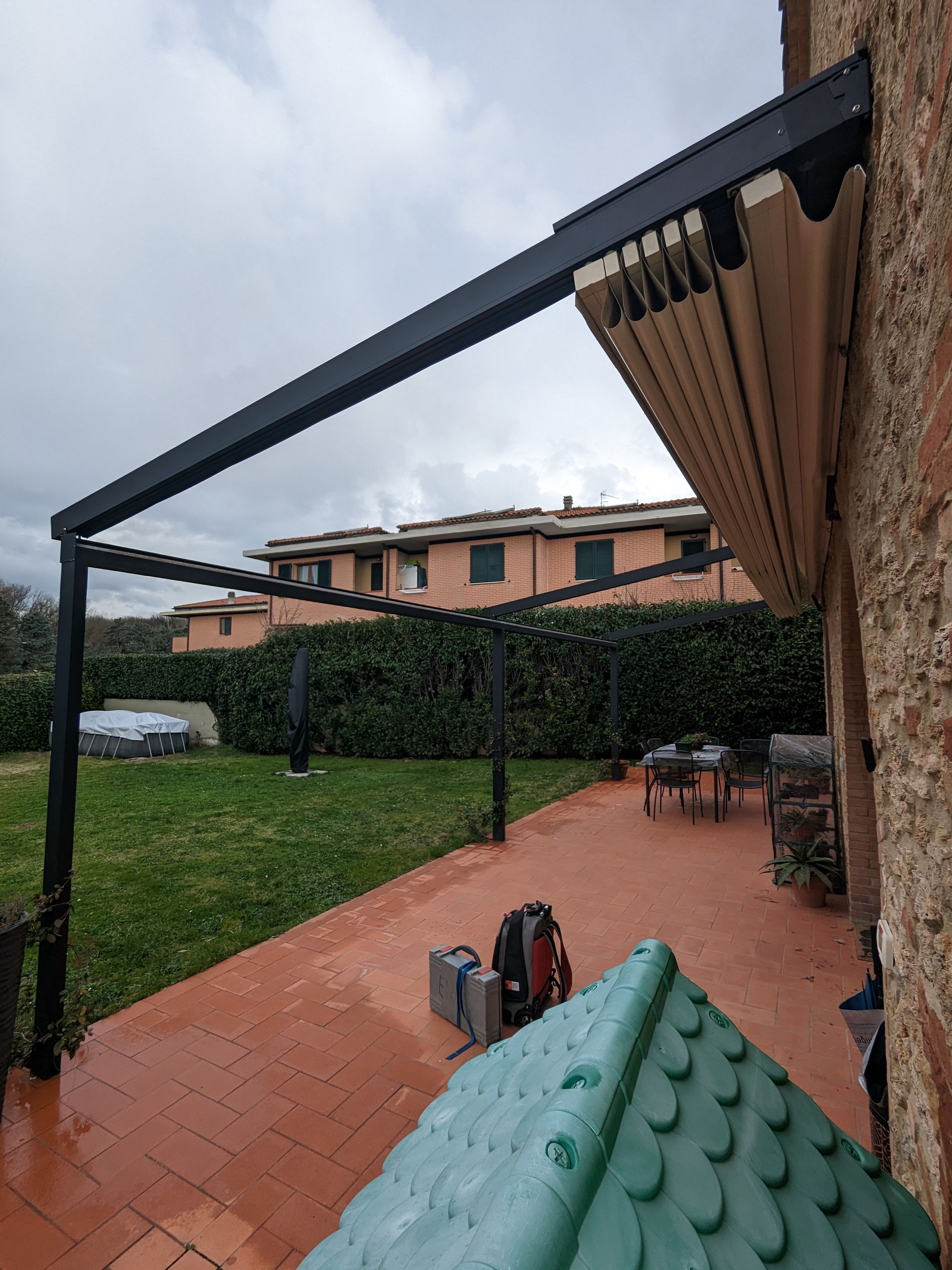 Patio coperto con pavimento in piastrelle rosse, struttura della tenda da sole nera e prato sul retro sotto un cielo nuvoloso.