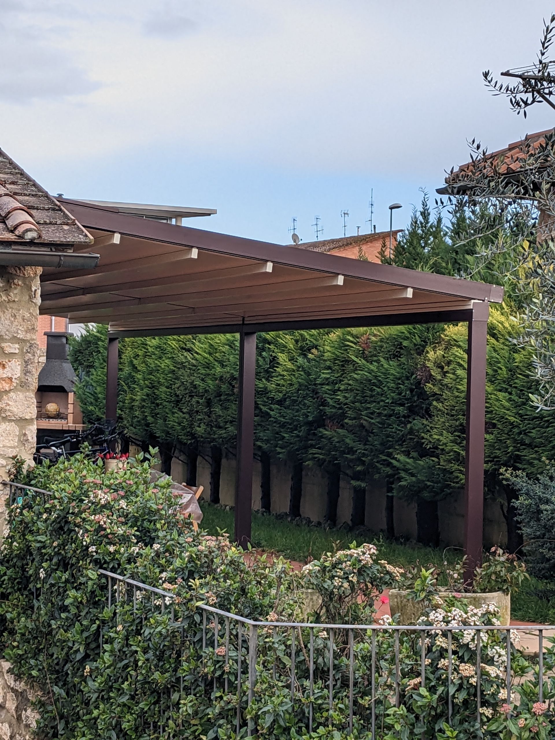 Pergola in legno sopra un sentiero del giardino accanto a una casa in pietra e a fitte siepi verdi sotto un cielo nuvoloso