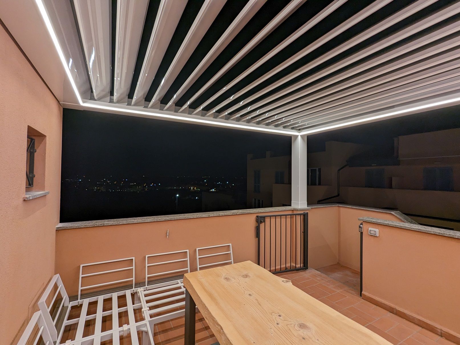 Terrazza coperta con tavolo in legno e sedie bianche con vista panoramica notturna.