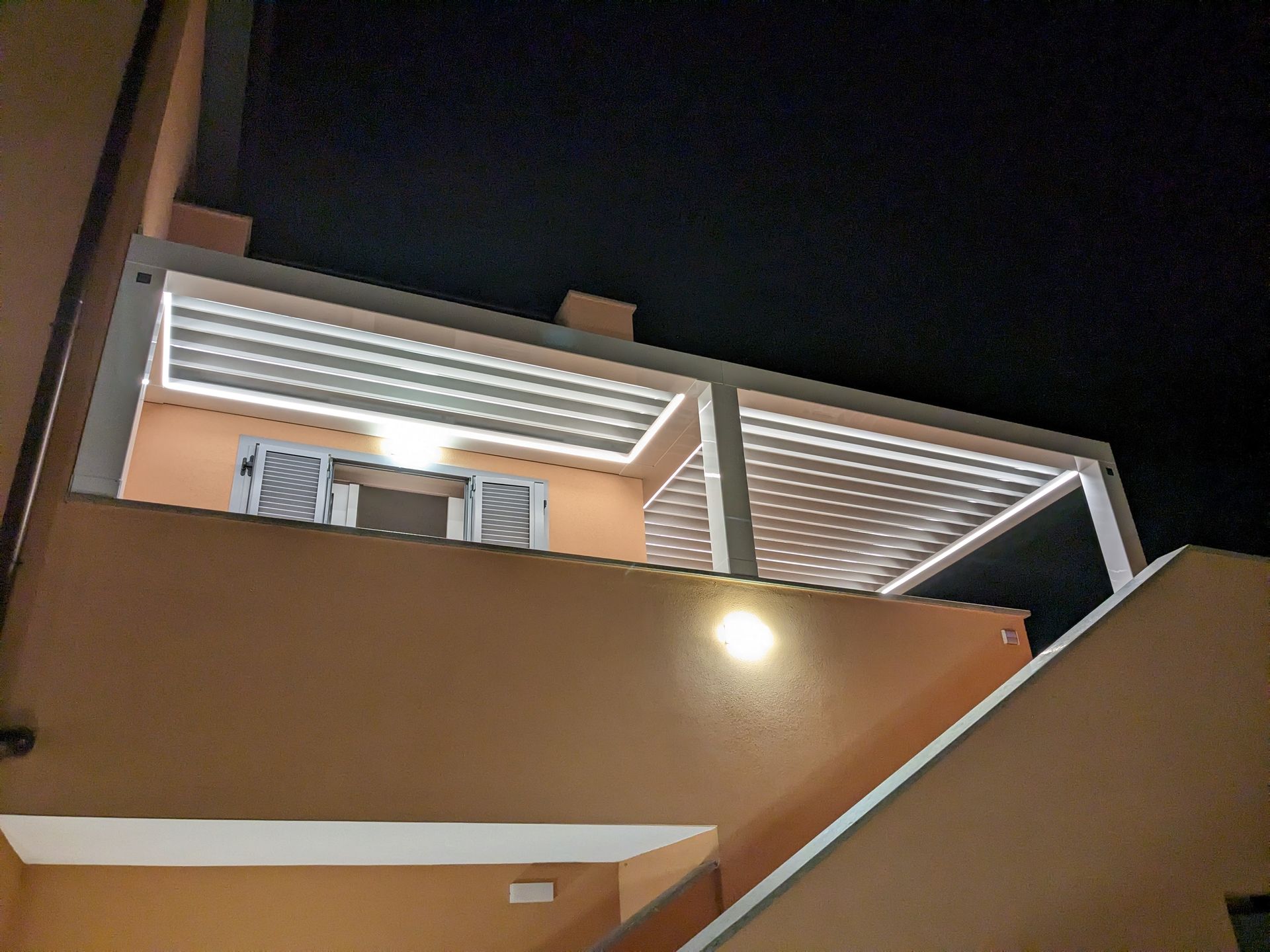Vista notturna del balcone di un edificio beige con luci brillanti e tende da sole sullo sfondo di un cielo scuro.