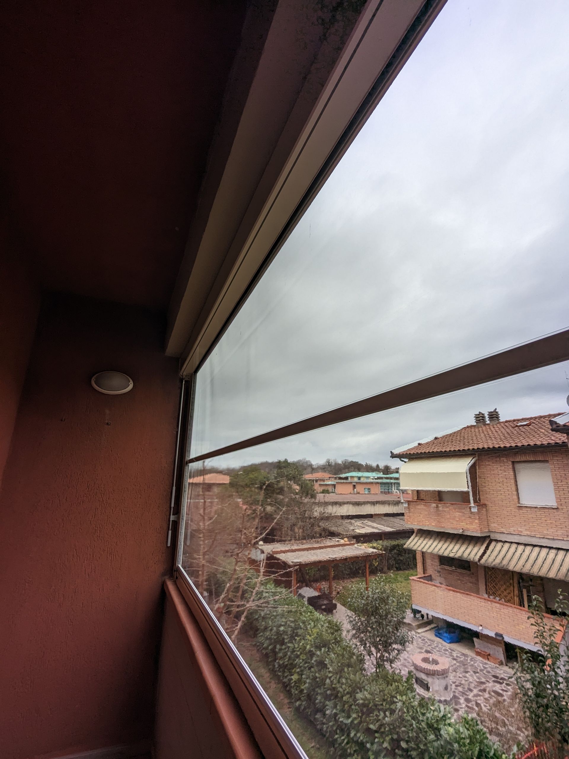 Vista dal balcone attraverso una finestra con zanzariera sui tetti vicini sotto un cielo nuvoloso.