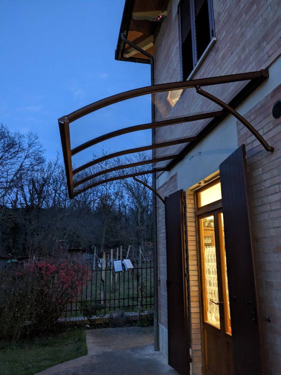 Esterno della casa al crepuscolo con finestra illuminata, persiana aperta e tenda da sole metallica curva sopra un patio.