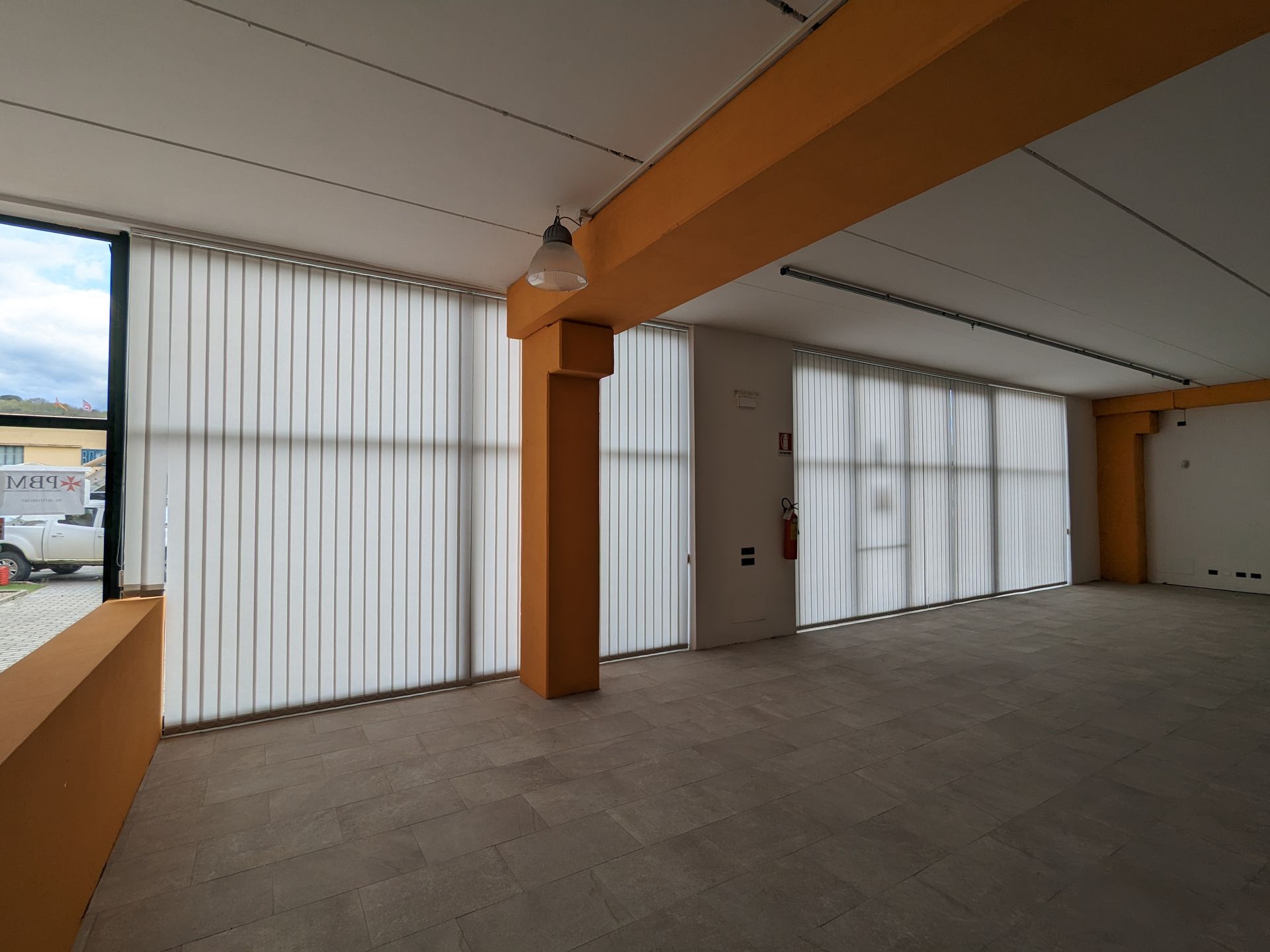 Stanza moderna vuota con soffitto inclinato, colonne arancioni e tende verticali bianche sopra le grandi finestre.