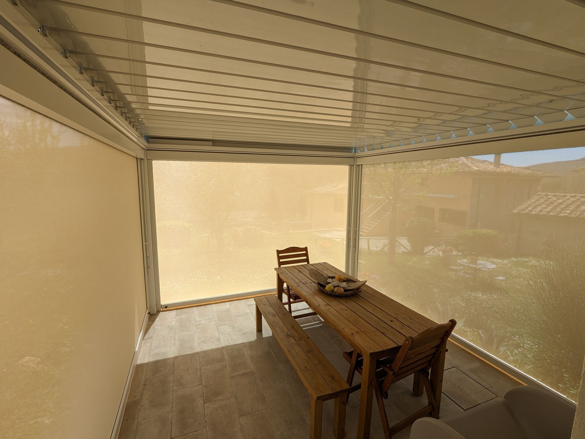 Patio ombreggiato con tavolo, panca e sedie in legno, accanto a una casa e a una rete da giardino.