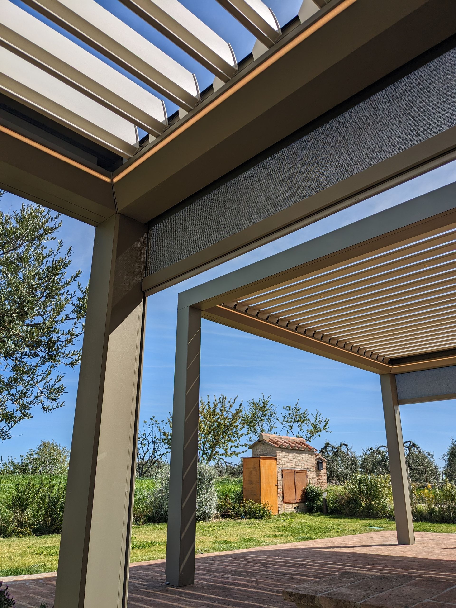Struttura moderna a pergola con tetto a doghe che si affaccia sul verde e su un piccolo edificio in lontananza.