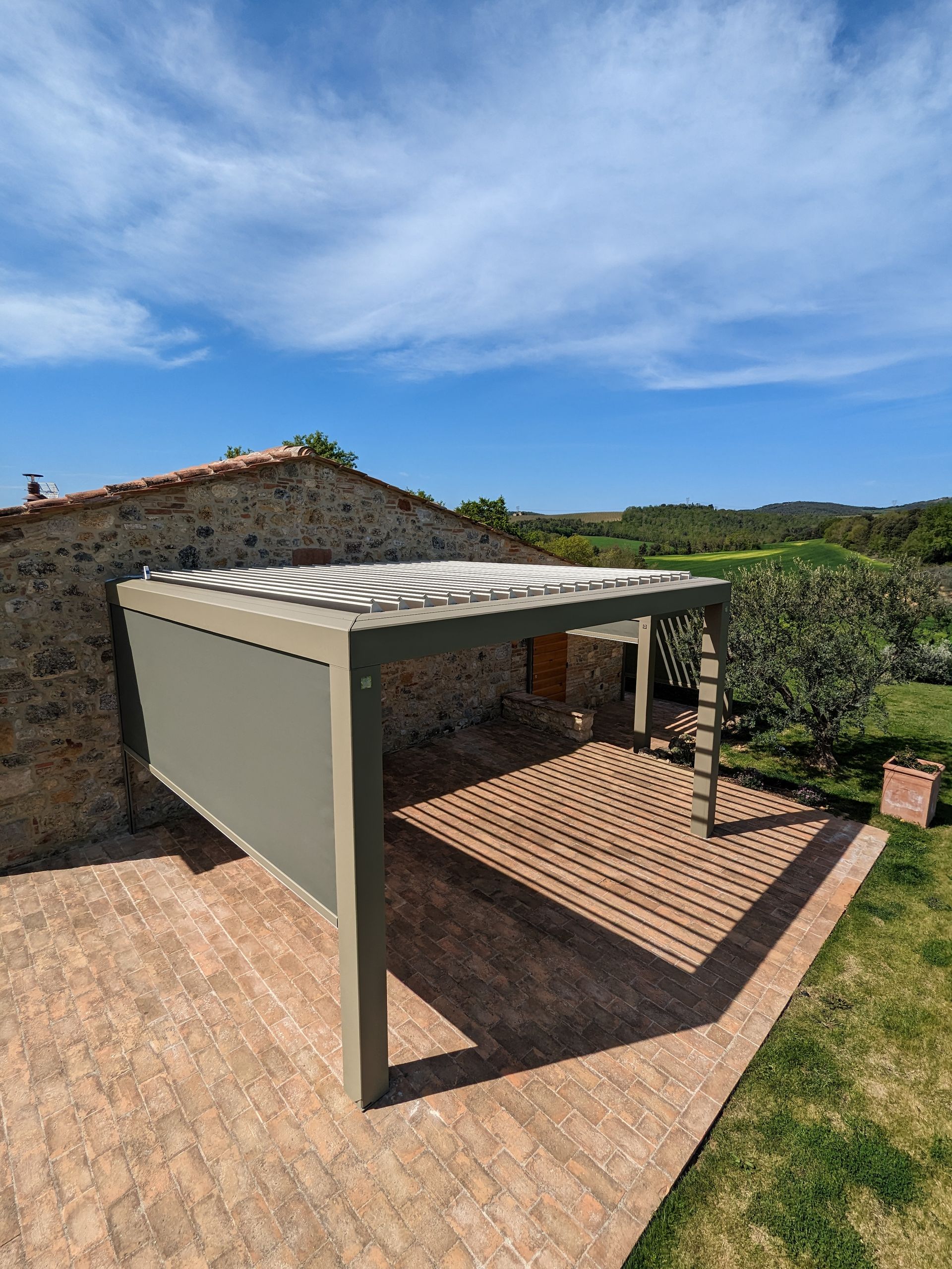 Piccolo patio esterno con pergolato accanto a un muro di pietra, sotto un cielo azzurro.