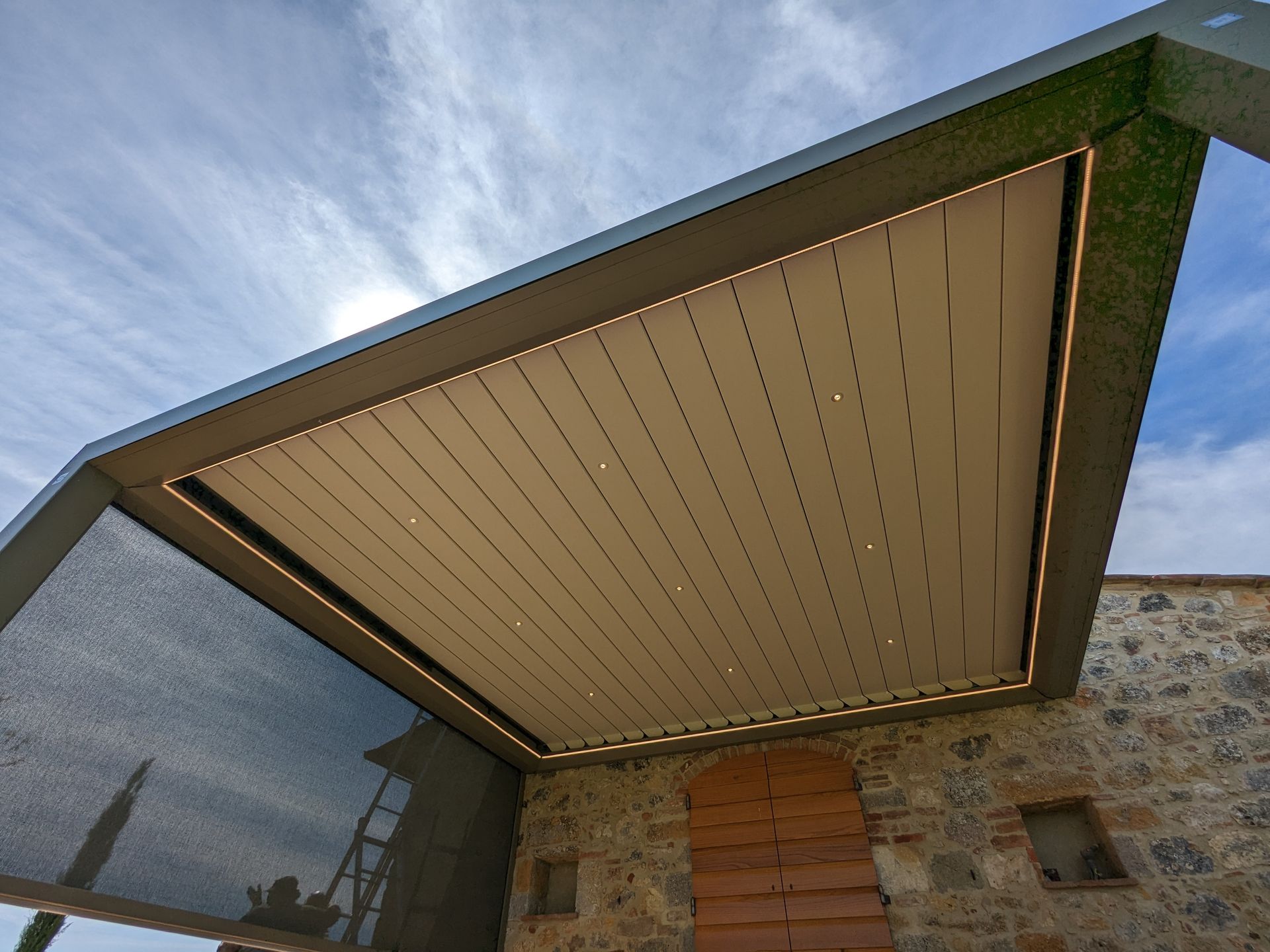 Ingresso di una casa moderna con una tettoia inclinata beige sopra un esterno in pietra e legno sotto un cielo azzurro
