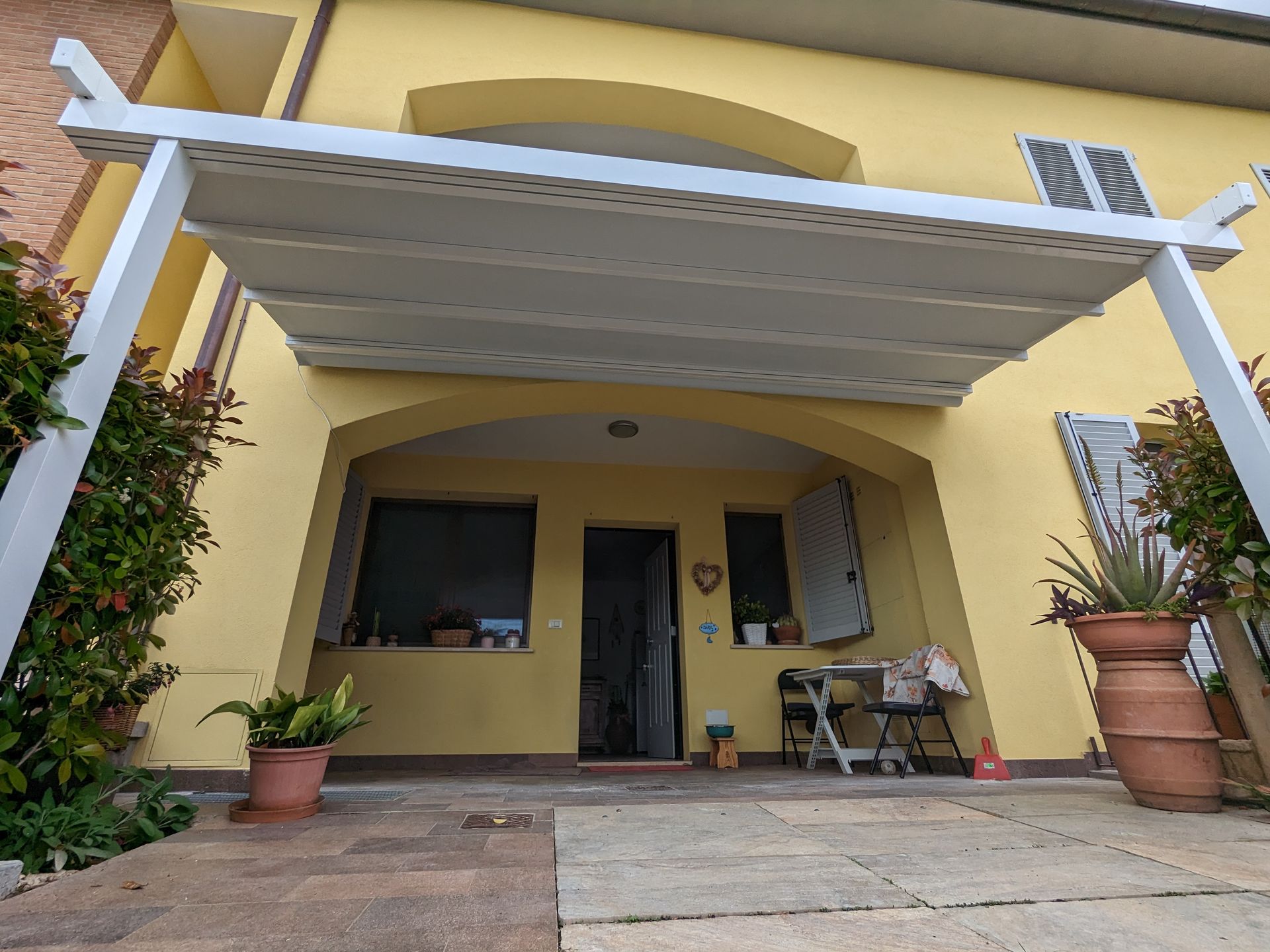 Veranda di una casa gialla con tenda da sole bianca, piante in vaso e sedie sotto l'ingresso coperto.