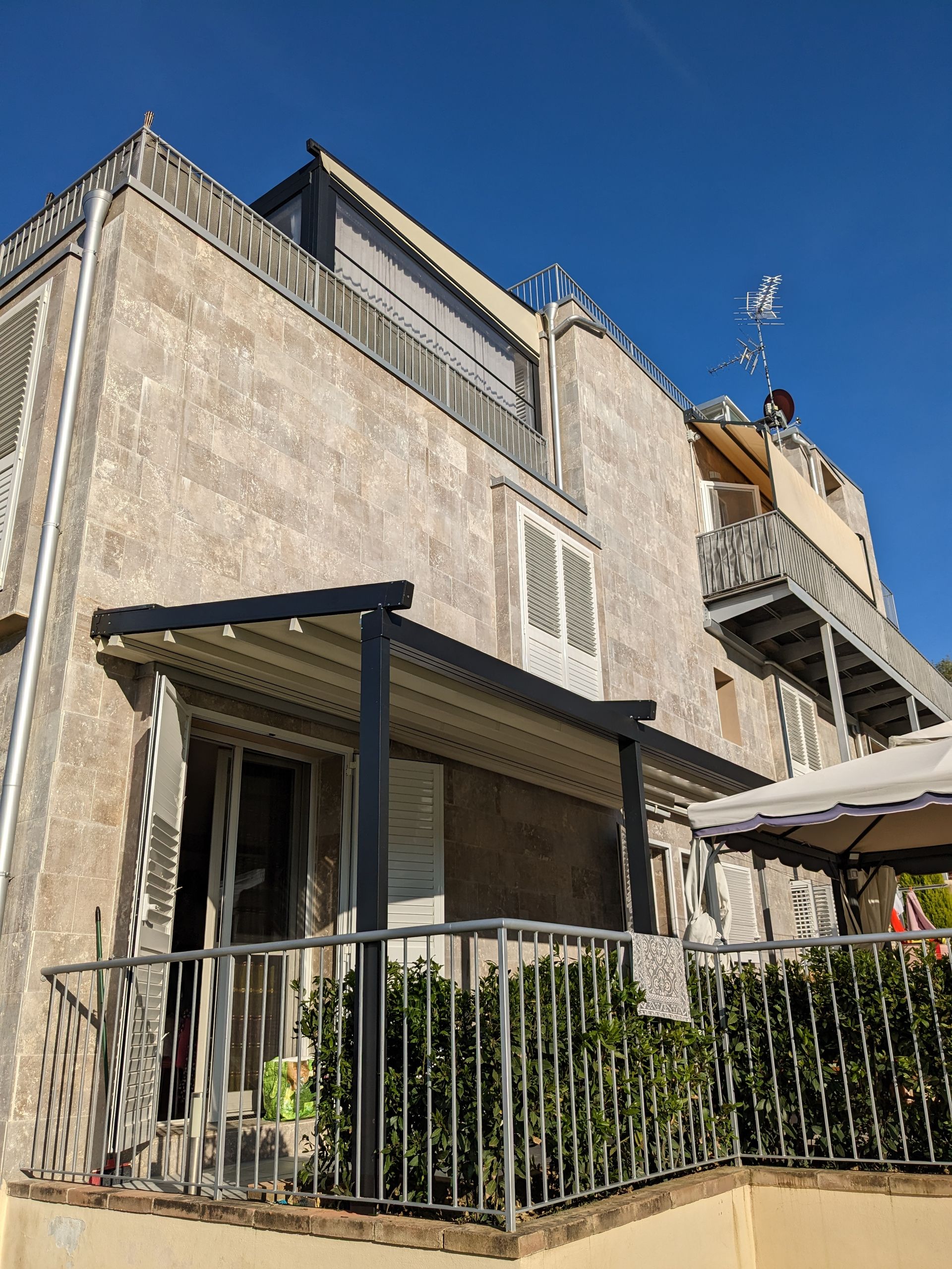 Facciata in pietra dell'edificio con balcone, ringhiera in ferro battuto e tenda da sole sotto un cielo azzurro limpido.