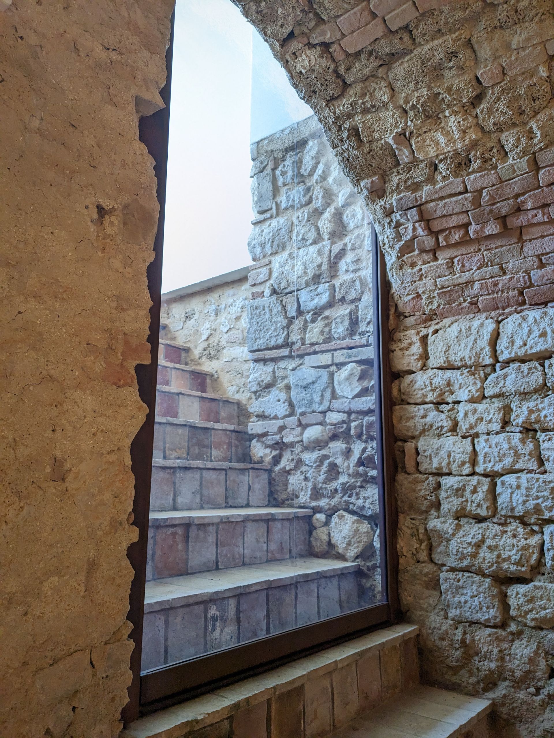 Scala in pietra vista attraverso una stretta finestra ad arco in un rustico muro di mattoni.