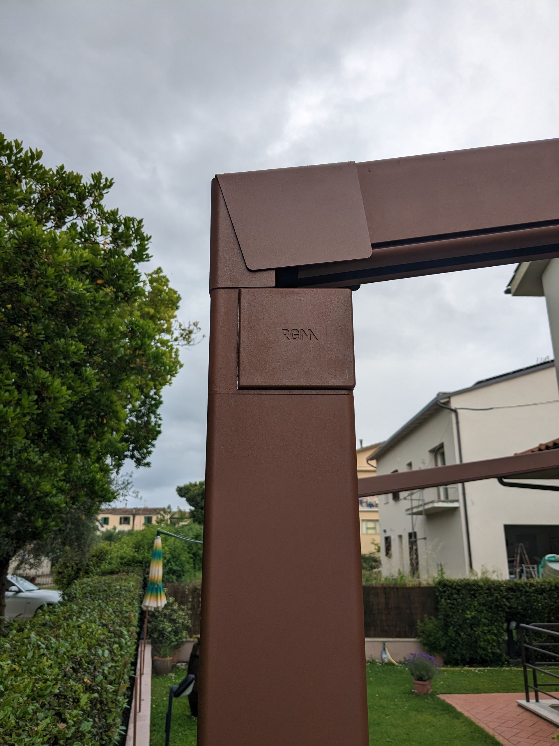 Un palo di sostegno marrone per la passerella si trova accanto a un cortile residenziale con siepi e un cielo nuvoloso.