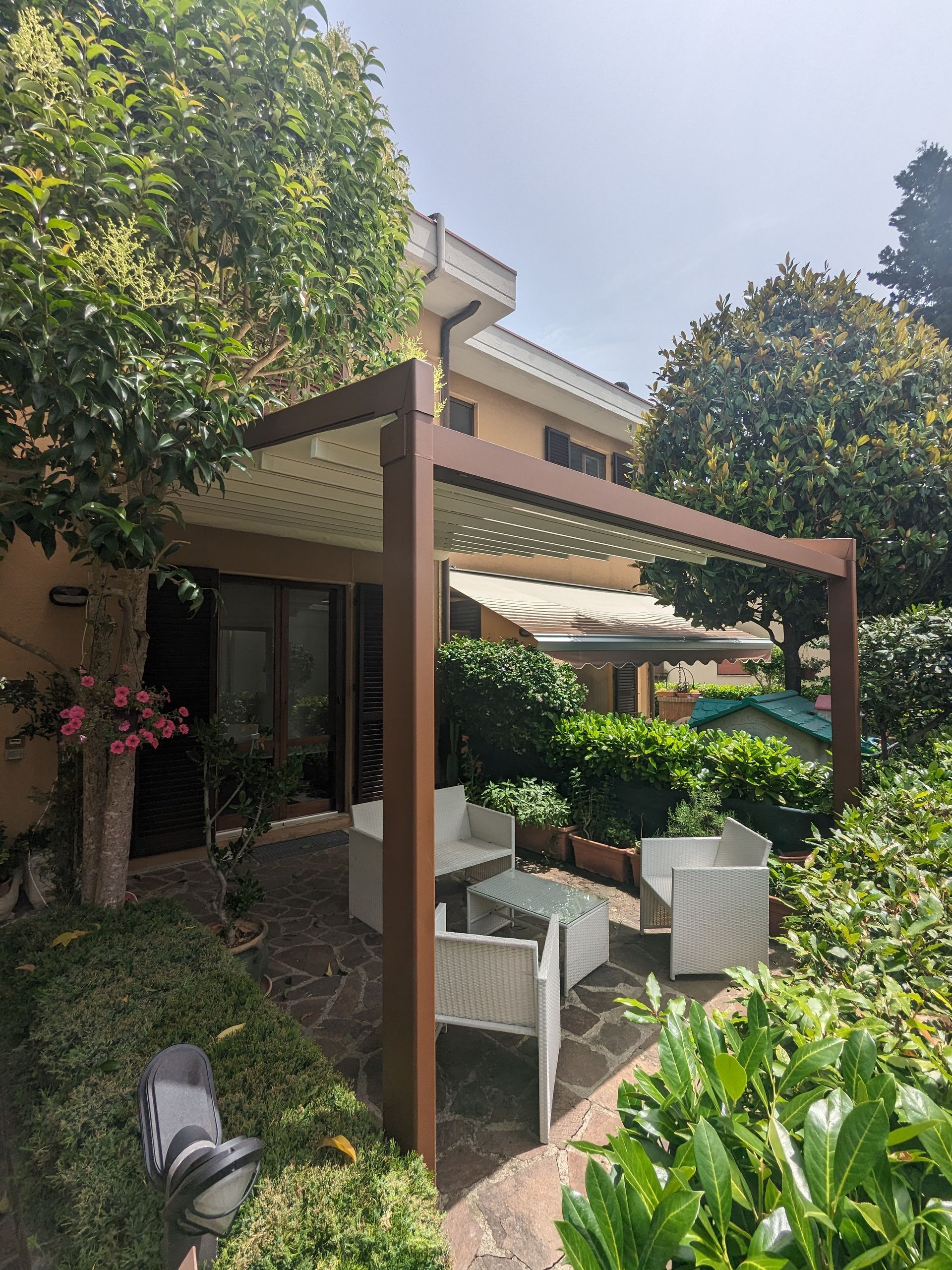Giardino sul patio con sedie bianche sotto un pergolato accanto a una casa, circondato da una vegetazione lussureggiante.