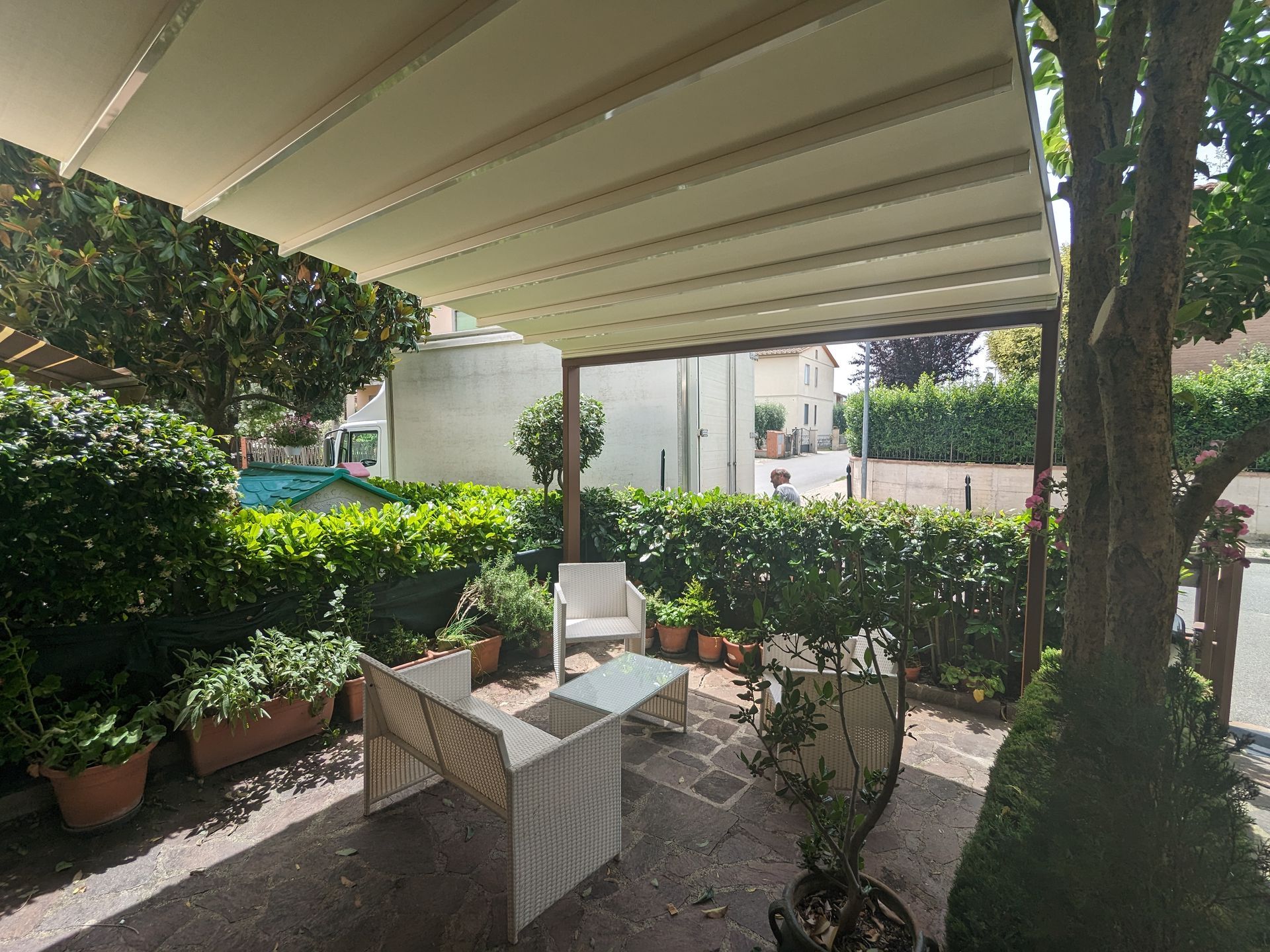 Patio coperto con sedie e tavolo in vimini, affacciato su un rigoglioso giardino e un cortile delimitato da siepi.