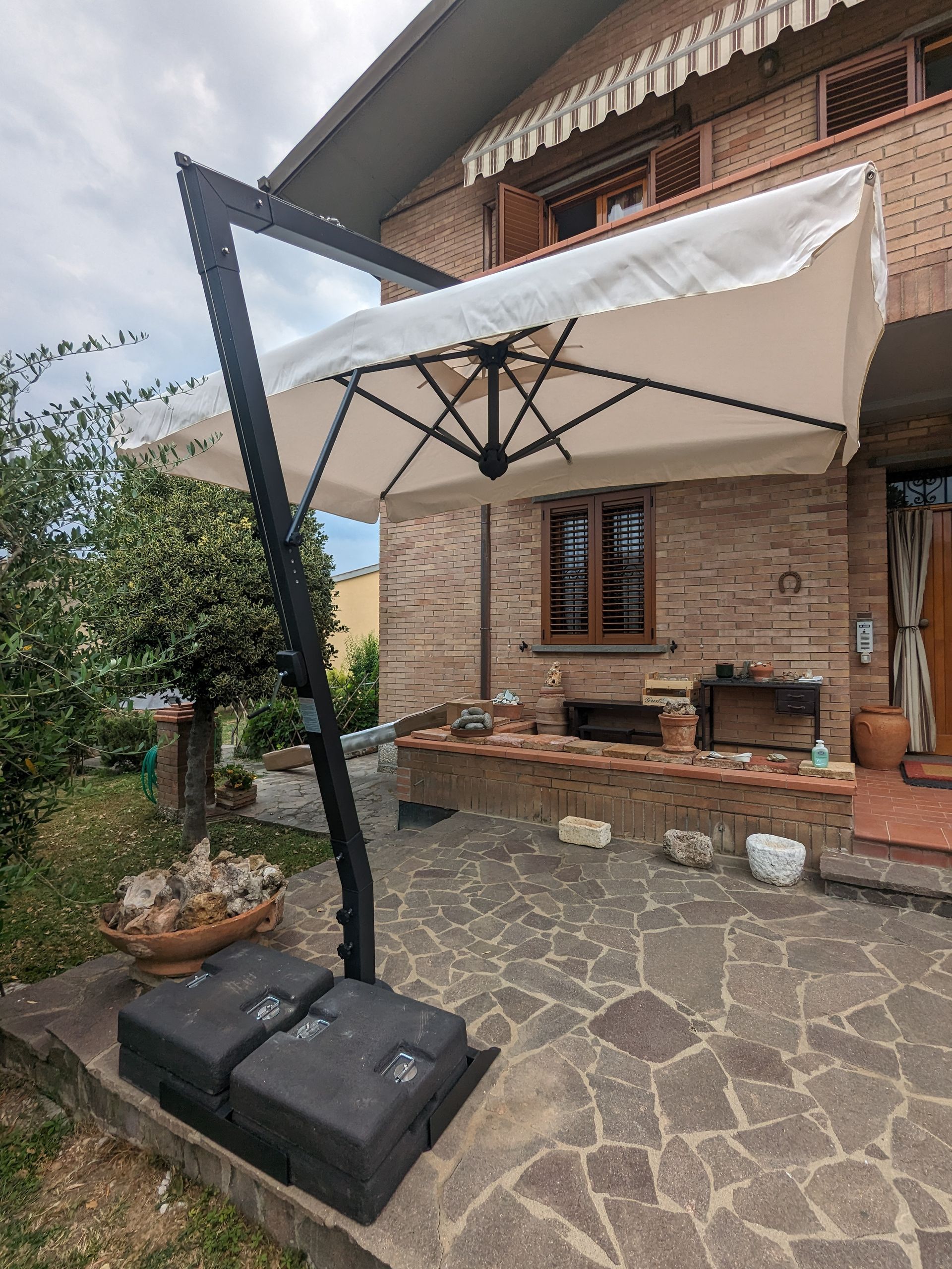 Ombrellone bianco da giardino sopra un patio in pietra accanto a una casa in mattoni e zona salotto esterna.