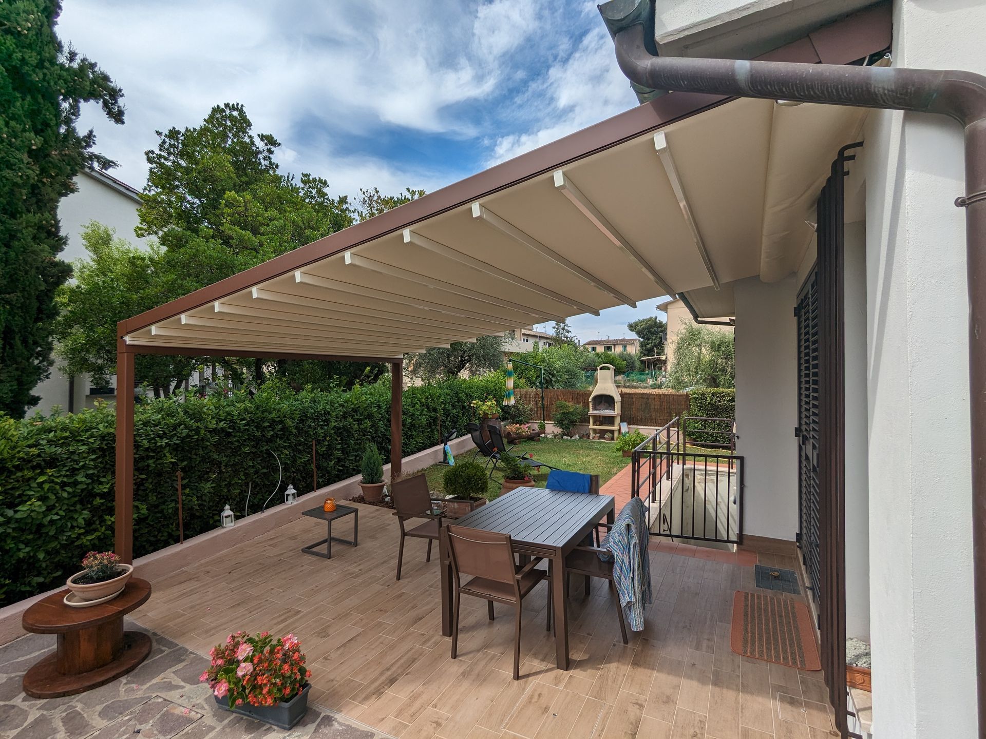 Patio posteriore con tenda da sole beige, tavolo e sedie da esterno, vasi di fiori e vista sul giardino.