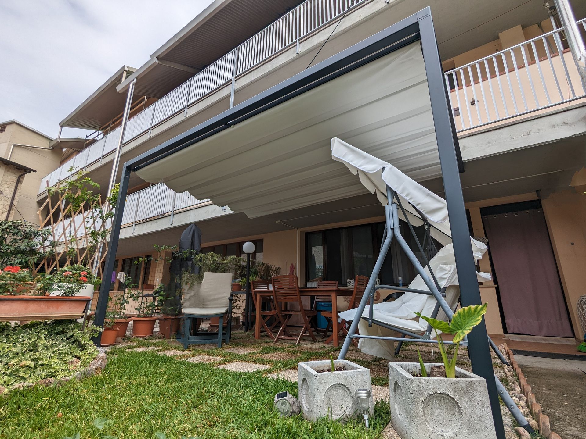 Patio sul retro della casa sotto una piattaforma rialzata con una sedia a dondolo sospesa, piante in vaso e un prato.