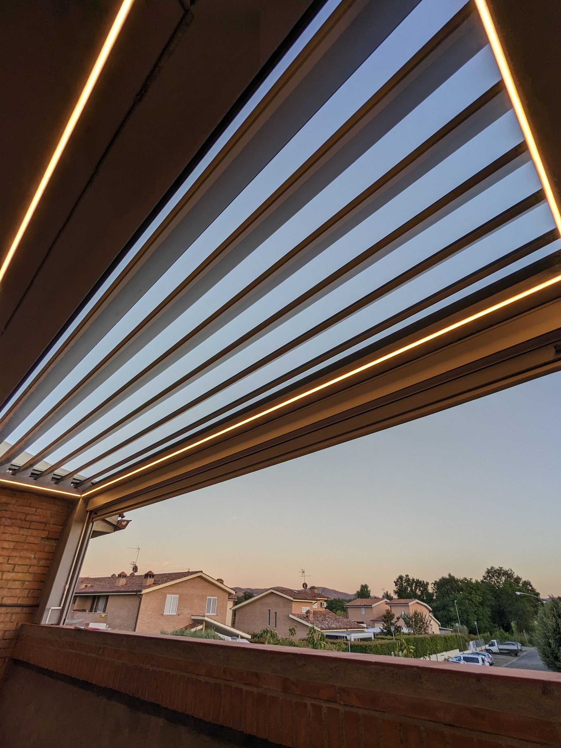 Tenda a pergola retrattile sopra un patio, che incornicia il cielo al tramonto e i tetti delle case di periferia.
