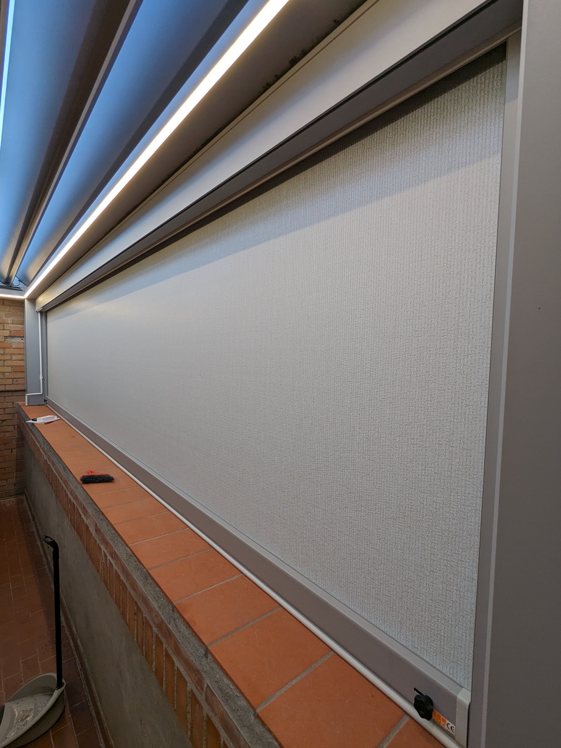 Balcone esterno con una lunga tenda a rullo bianca e davanzale rivestito in piastrelle di terracotta lungo la parete.