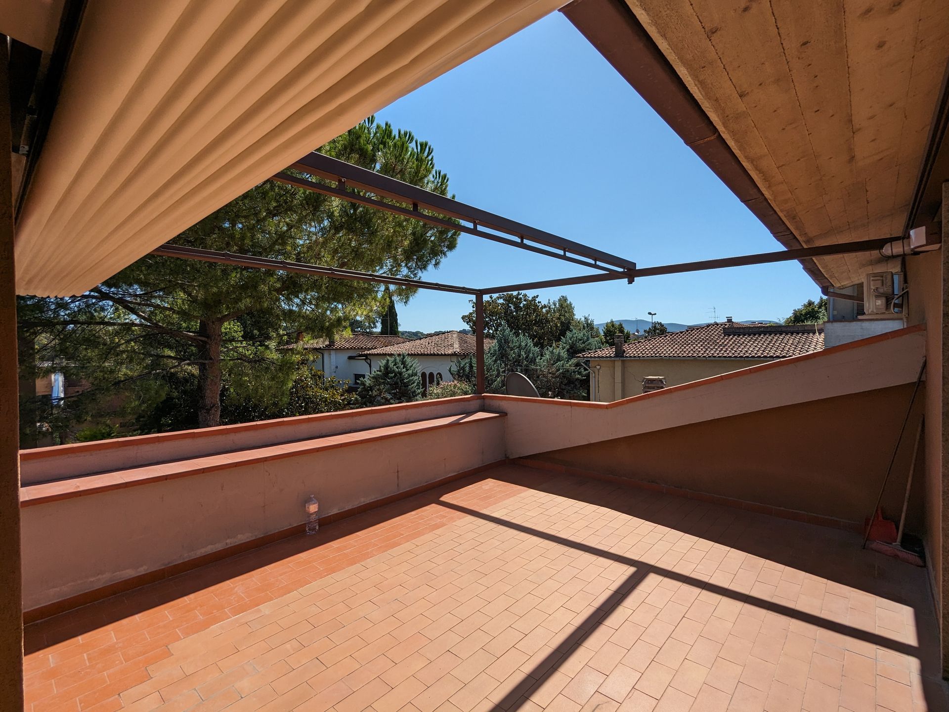 Balcone soleggiato in terracotta con tenda da sole retrattile, muretto basso e alberi oltre la ringhiera.