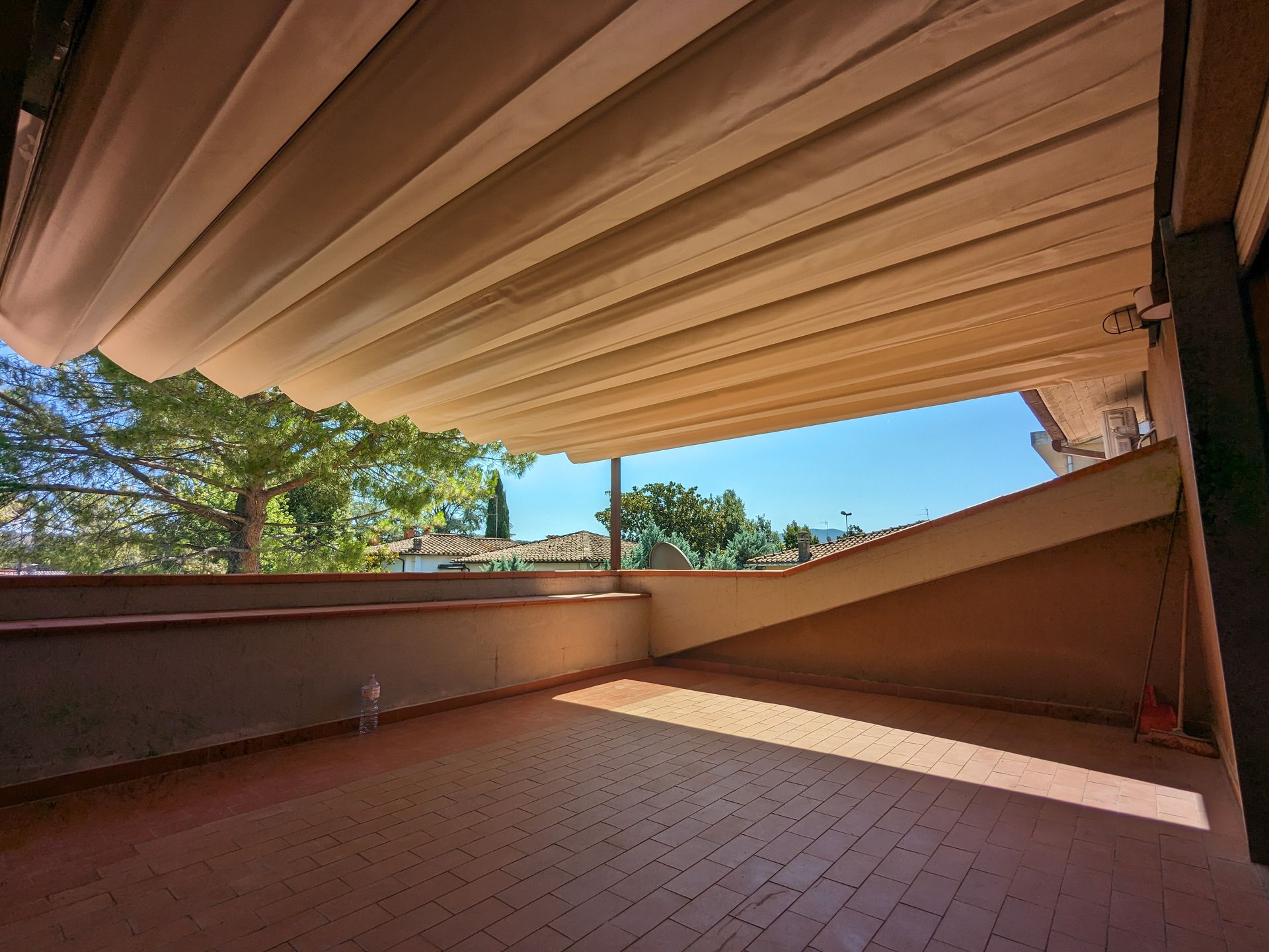 Patio coperto con tenda da sole retrattile a lamelle, pavimento in piastrelle di terracotta e alberi visibili oltre la ringhiera.