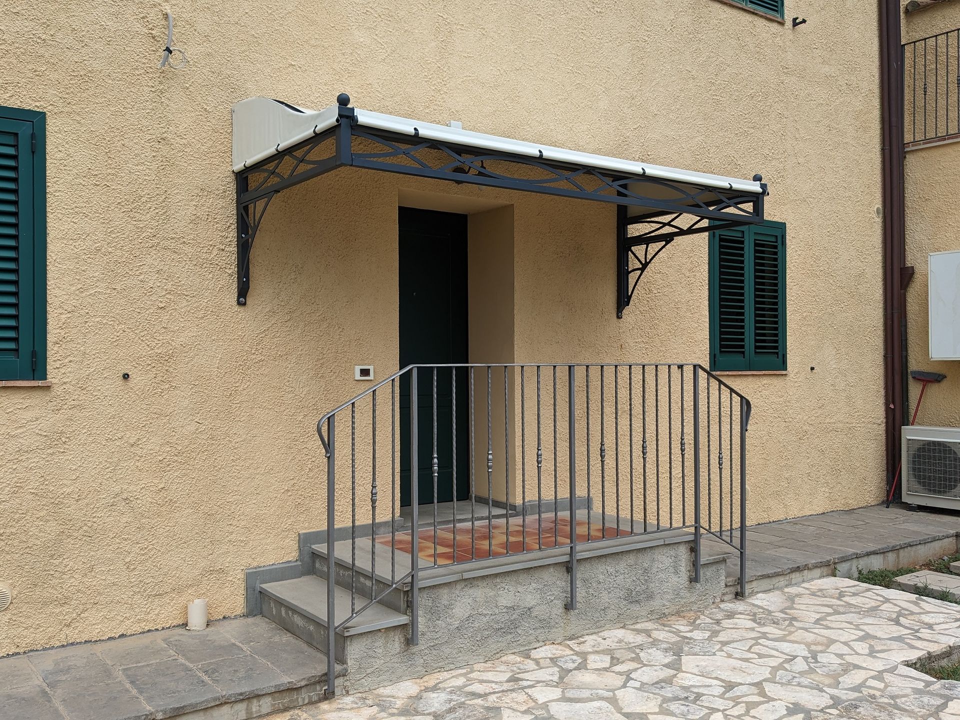 Piccolo ingresso di casa beige con tettoia in metallo, gradini e rampa accanto a persiane verdi