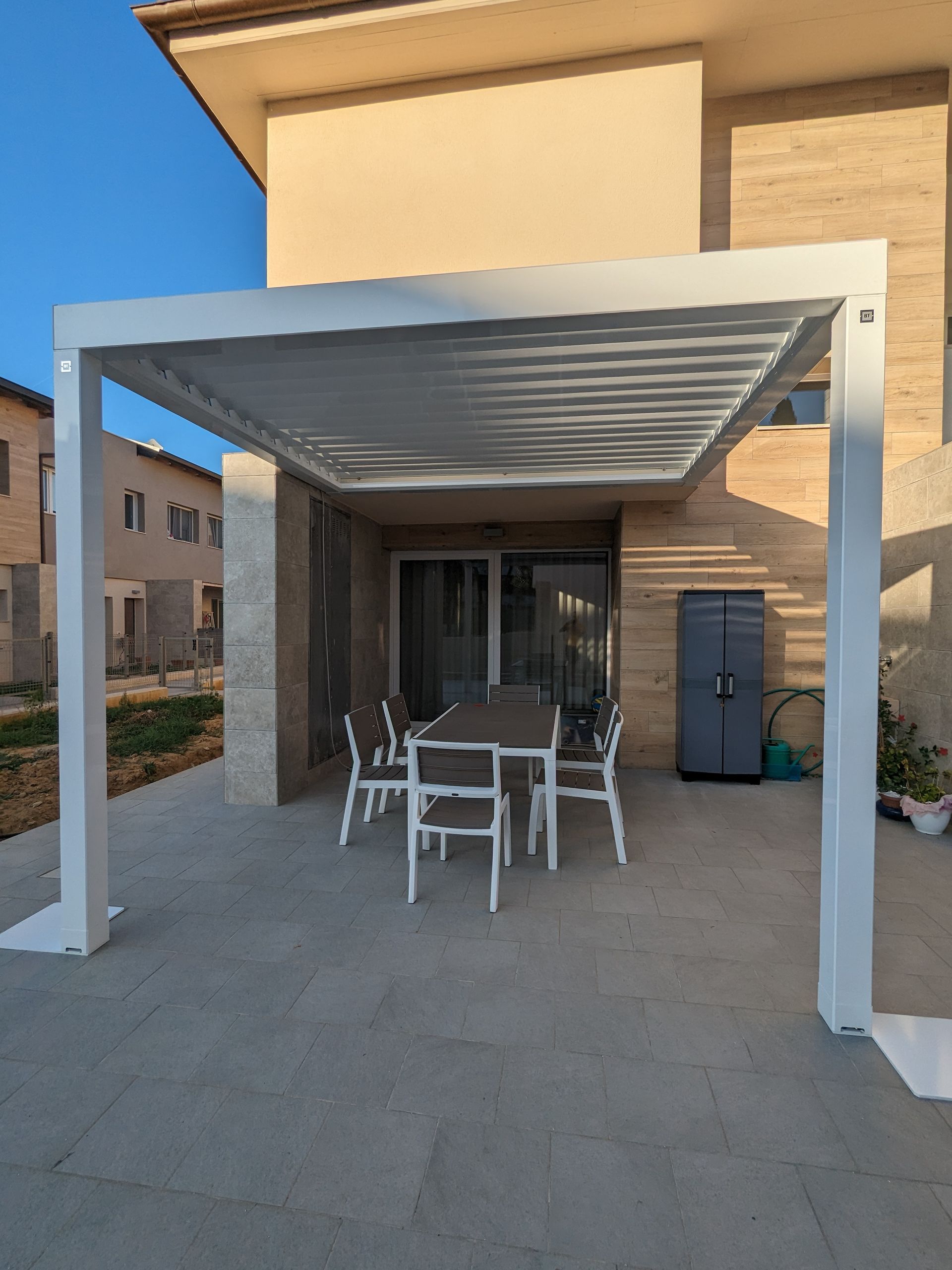 Patio esterno con tavolo e sedie bianchi sotto un pergolato moderno accanto all'ingresso di un edificio.