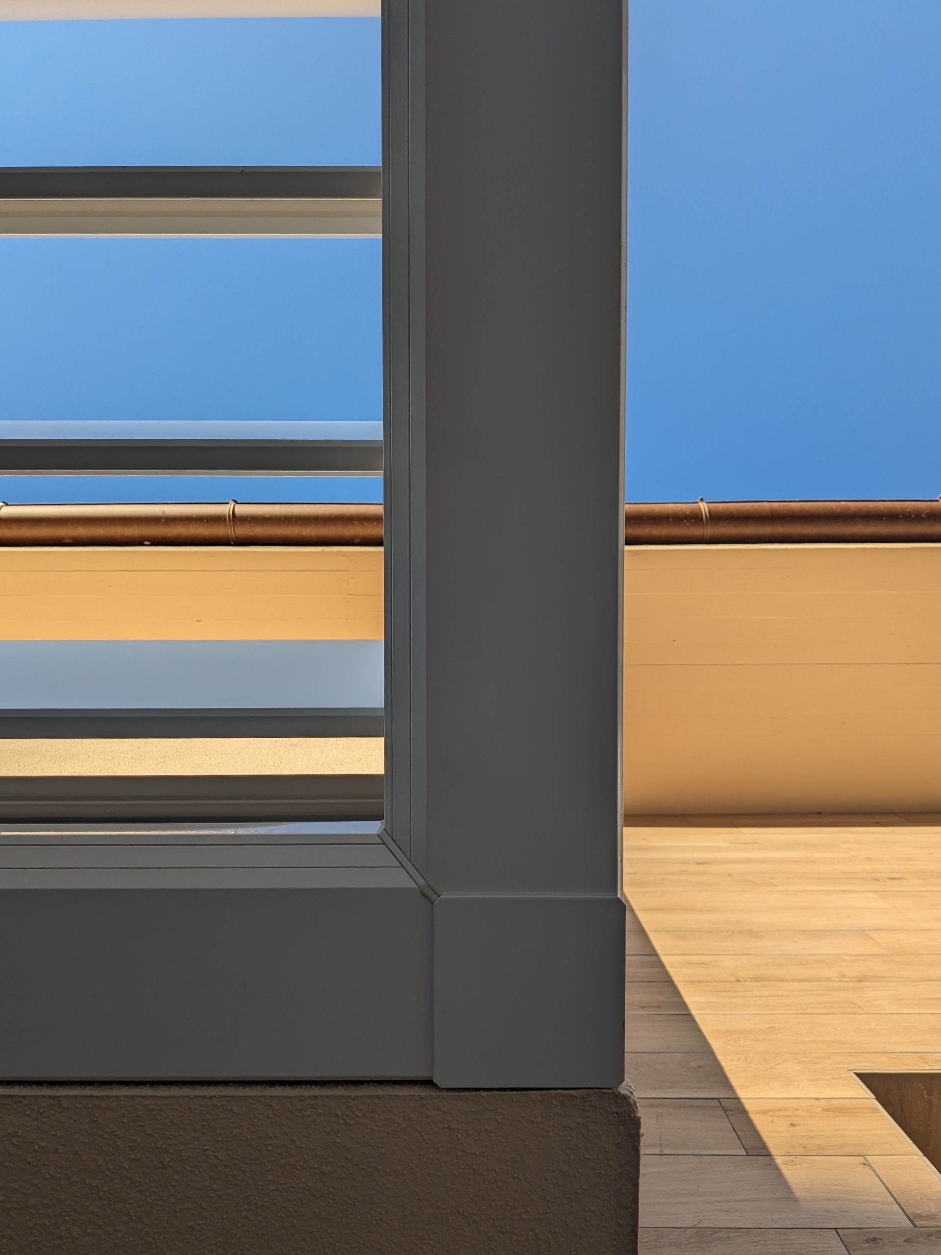 Colonna grigia accanto a una finestra con cielo azzurro e parete beige, accanto a un pavimento in legno chiaro e un tappeto scuro.