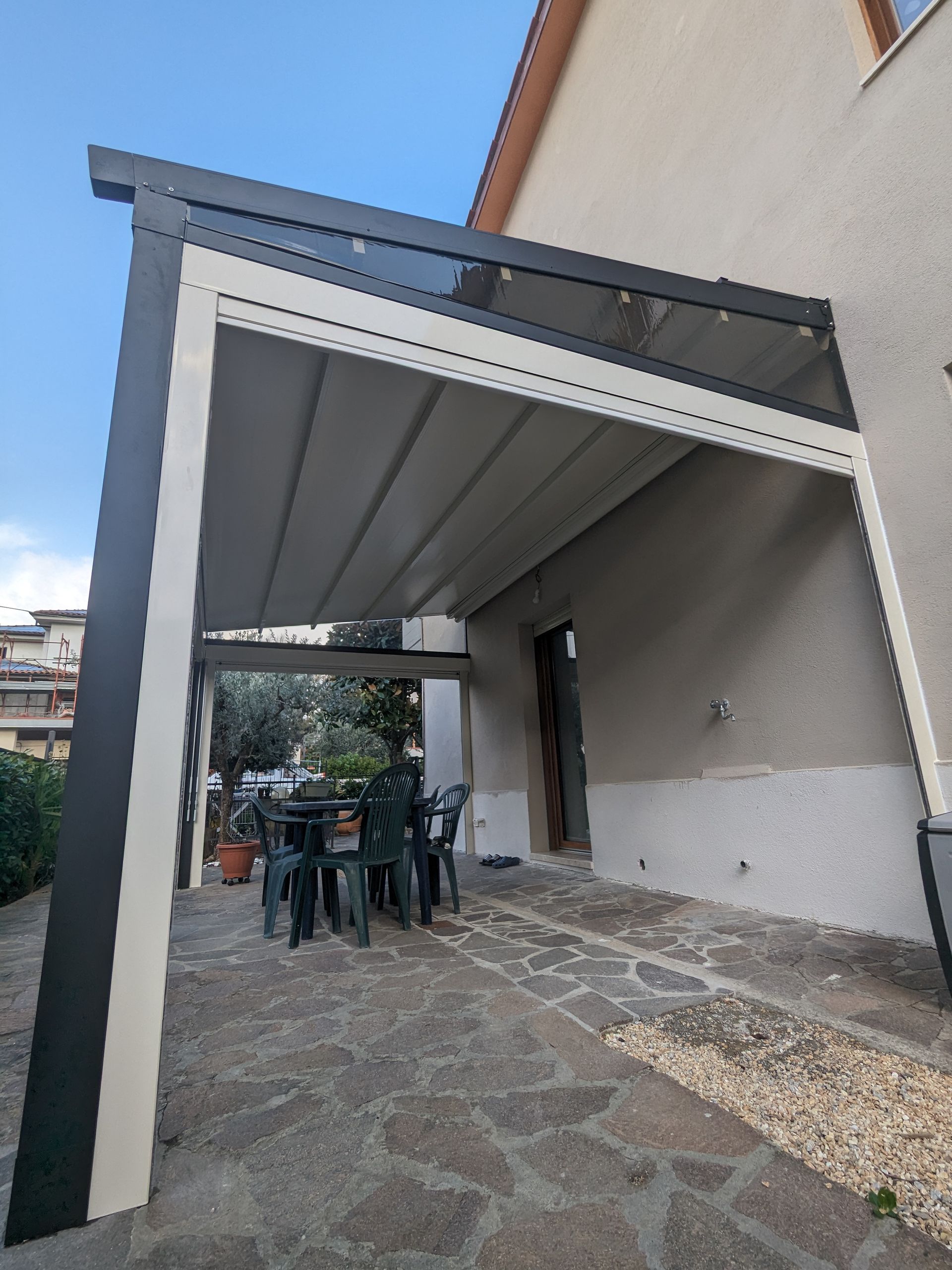 Patio esterno coperto con pavimento in pietra, tettoia in metallo e persone sedute a un tavolo vicino a un edificio.