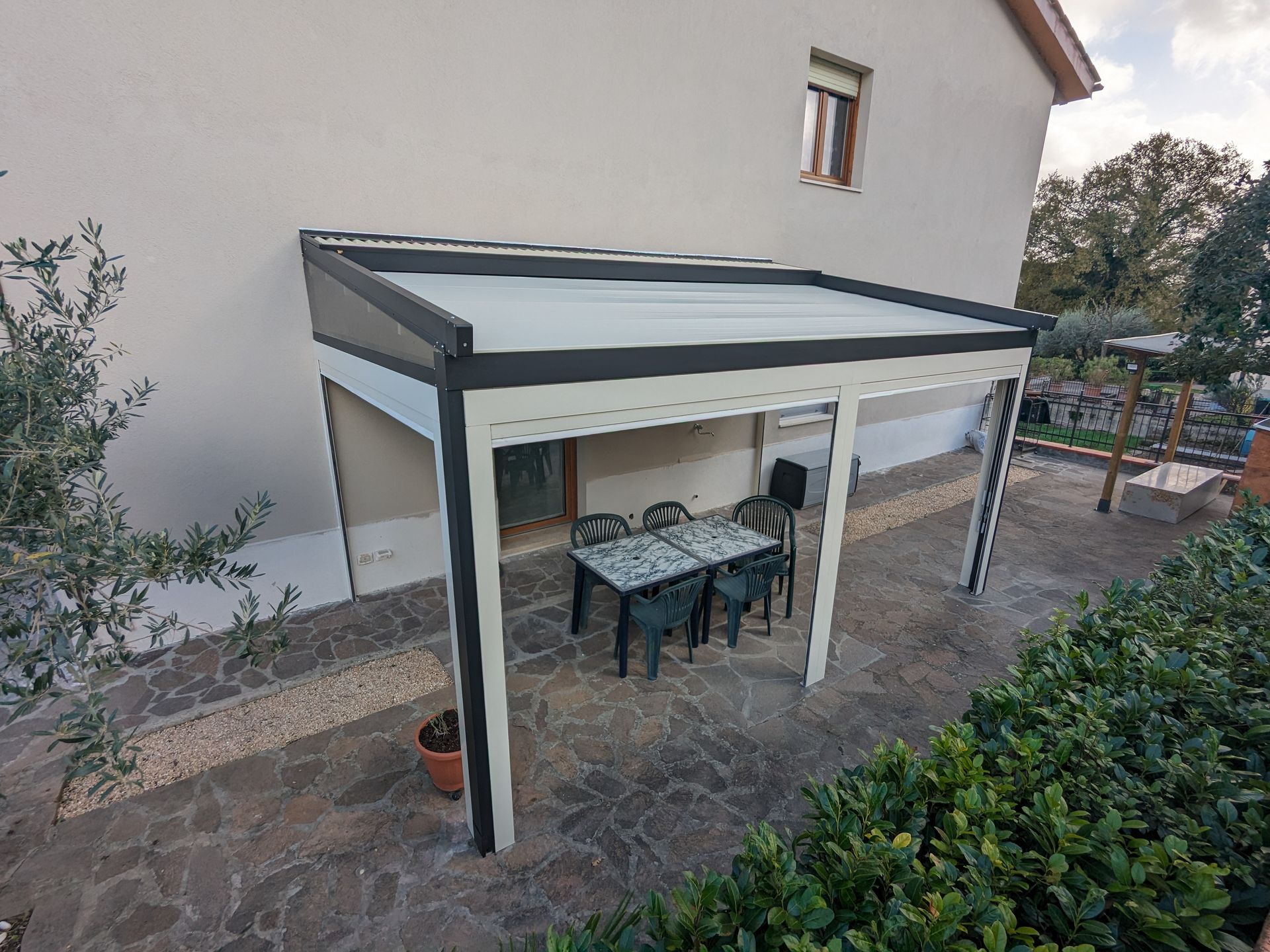 Patio esterno con tenda da sole bianca e nera, tavolo da pranzo e sedie, adiacente a una casa.