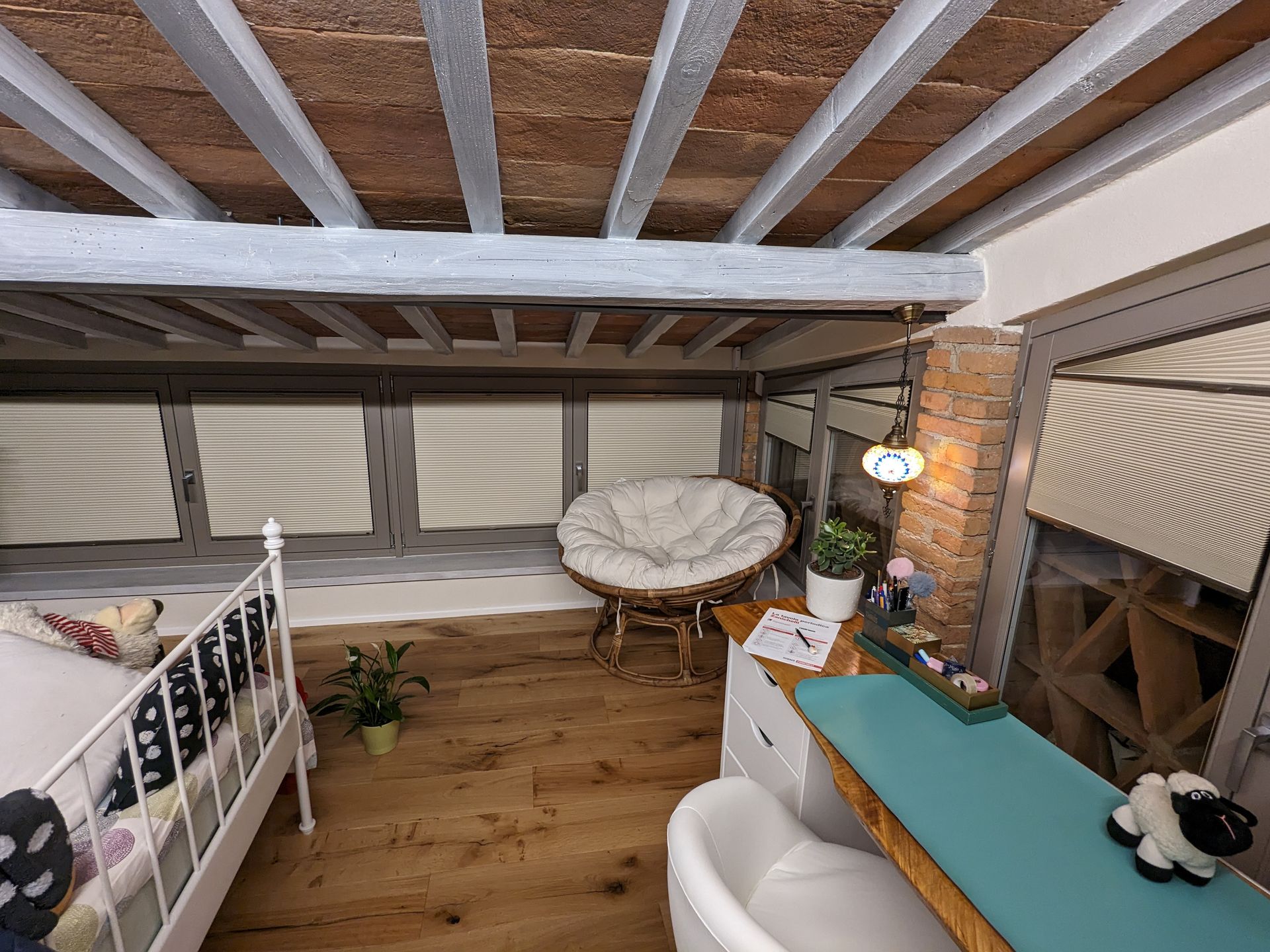Piccola camera da letto mansardata con ringhiera bianca, travi in legno, divano letto, sedia, scrivania e lucernario.