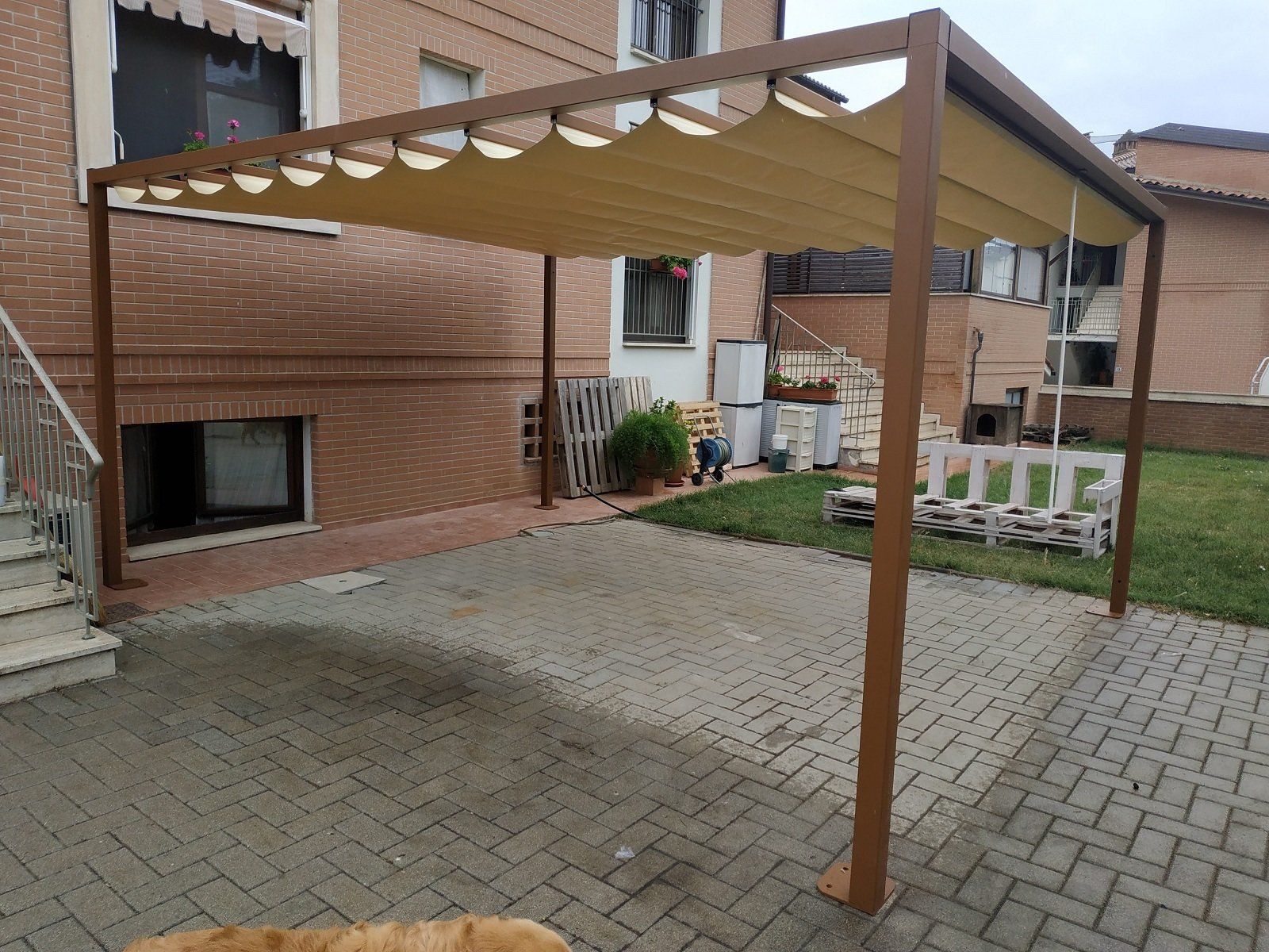 Pergola marrone da patio con tettoia color beige sopra un cortile pavimentato accanto a un edificio in mattoni.