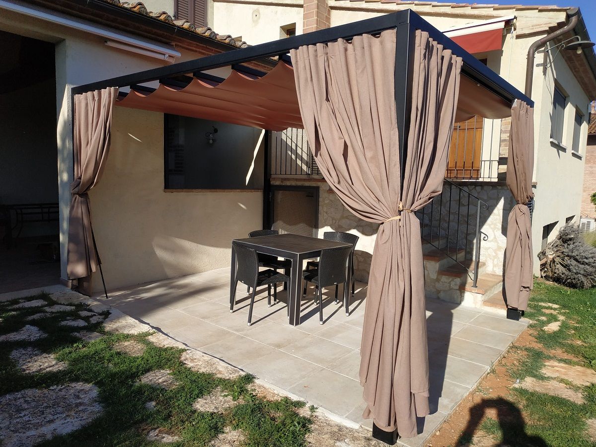 Patio coperto con tende beige, tavolo e sedie da esterno su un patio in cemento accanto a una casa.