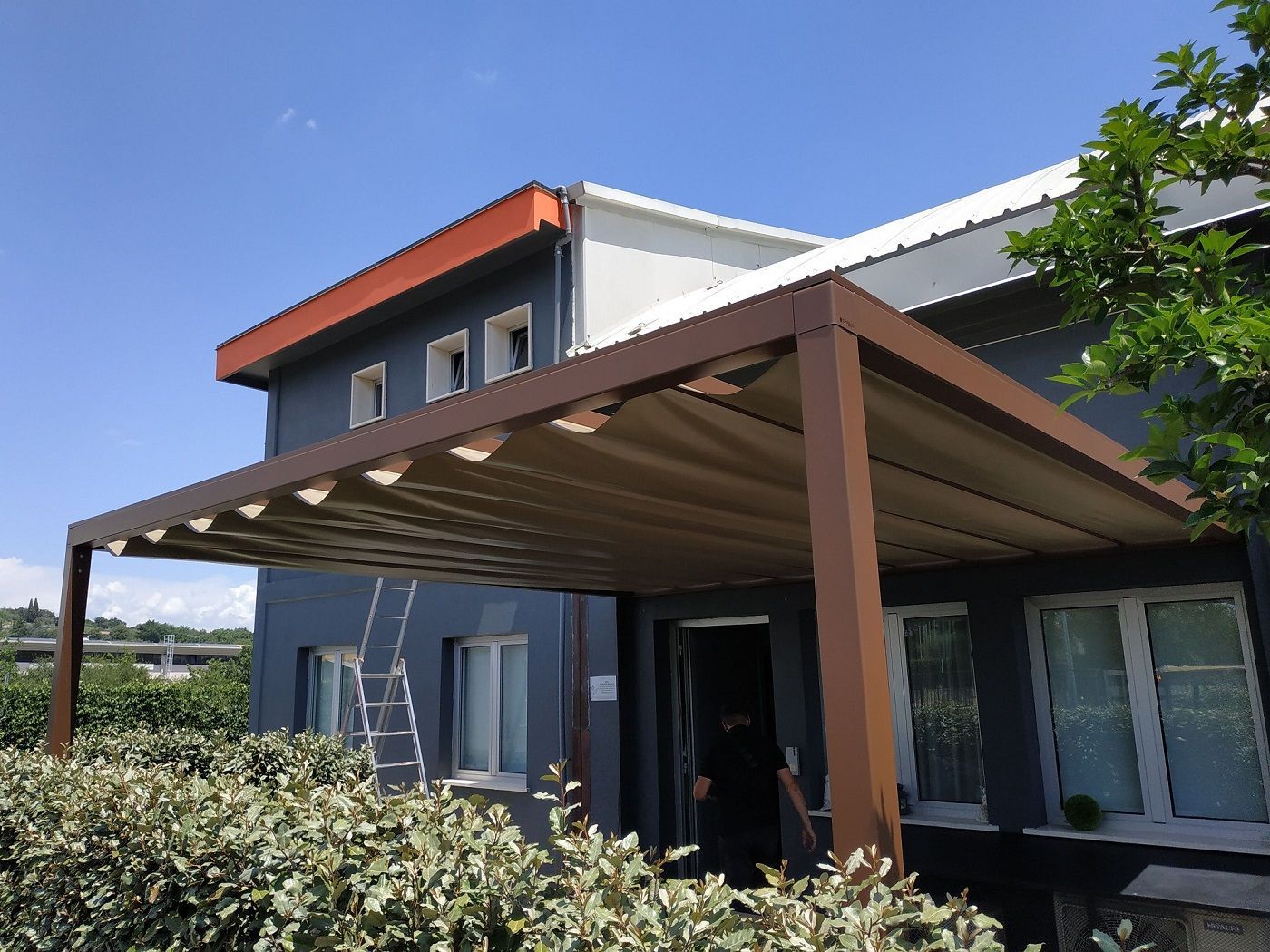 Casa moderna a due piani con tettoia marrone, scala e siepe sotto un cielo azzurro limpido.