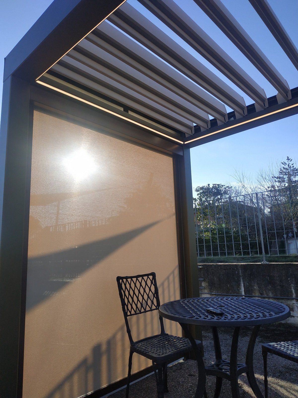 Patio con tavolo e sedia in metallo nero accanto a un muro beige sotto un pergolato a doghe al tramonto