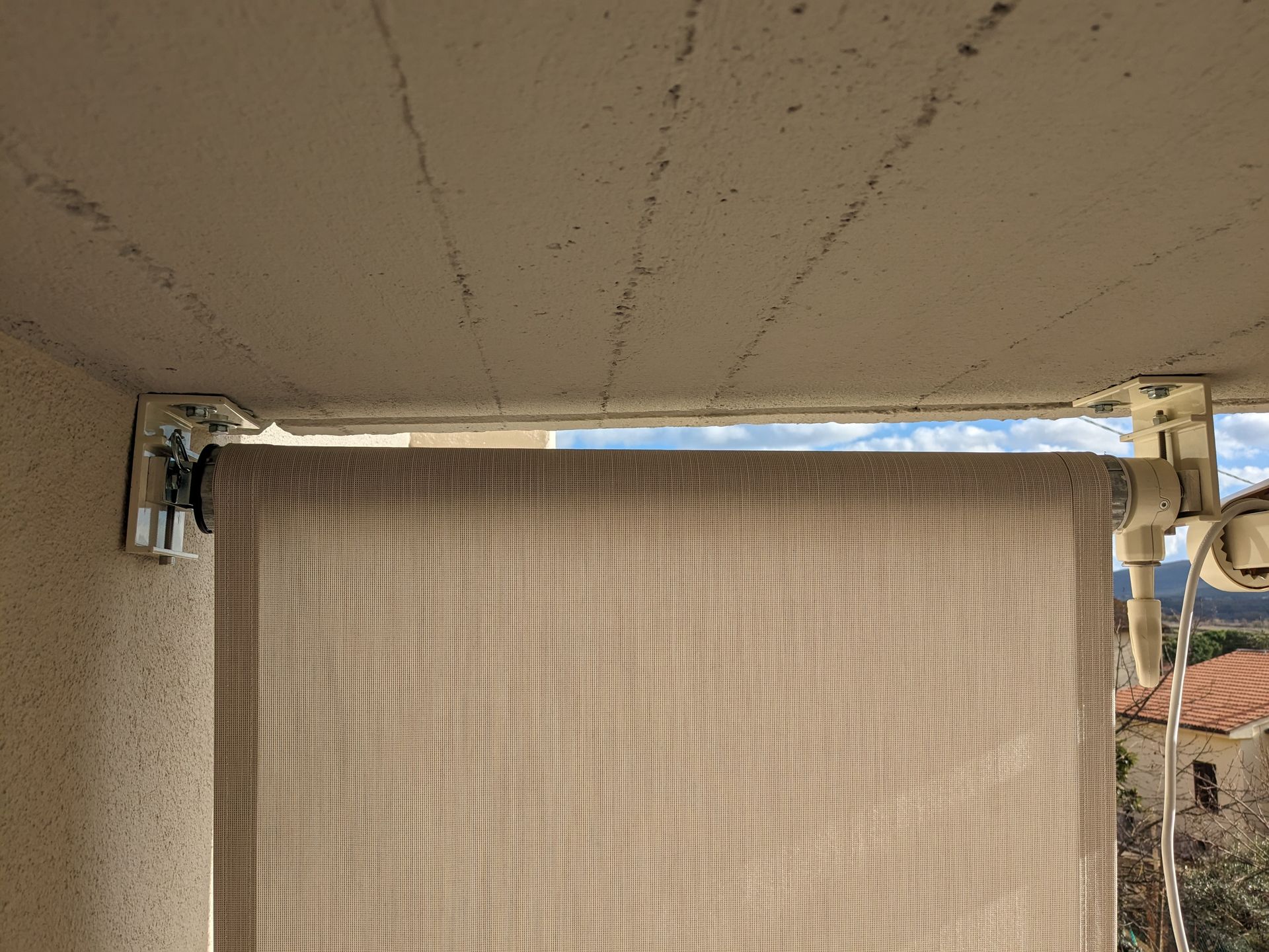 Tenda da sole beige chiusa sotto una tettoia in cemento, con tegole del tetto visibili sul bordo destro.