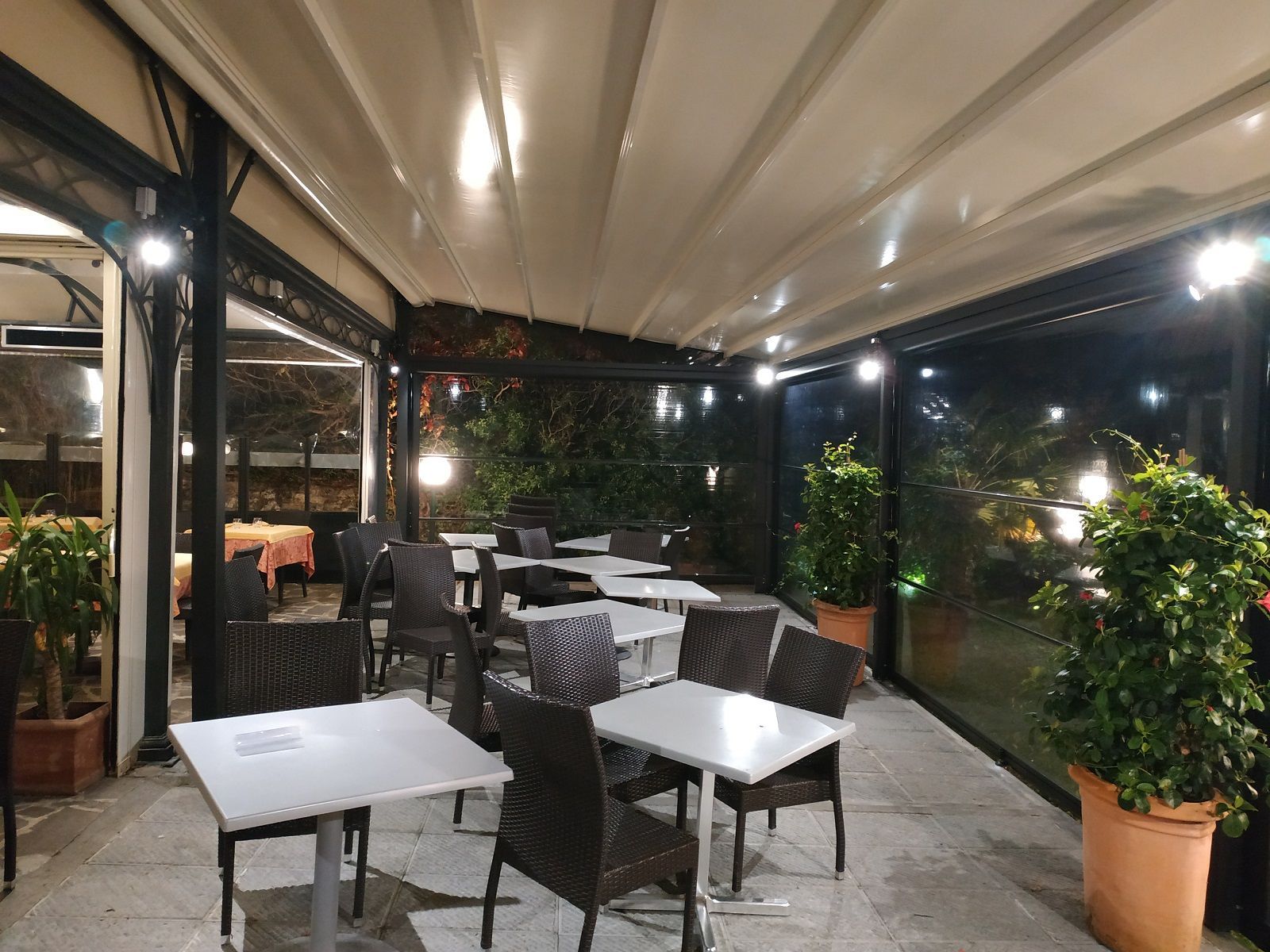 Tavolini vuoti all'aperto di un caffè, sotto una tettoia, di notte, con tavolini bianchi, sedie scure e piante in vaso.