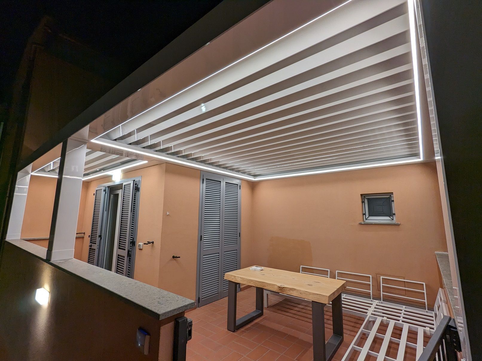 Patio coperto con tavolo in legno, sedie bianche, pareti color pesca e soffitto a pergola con doghe.