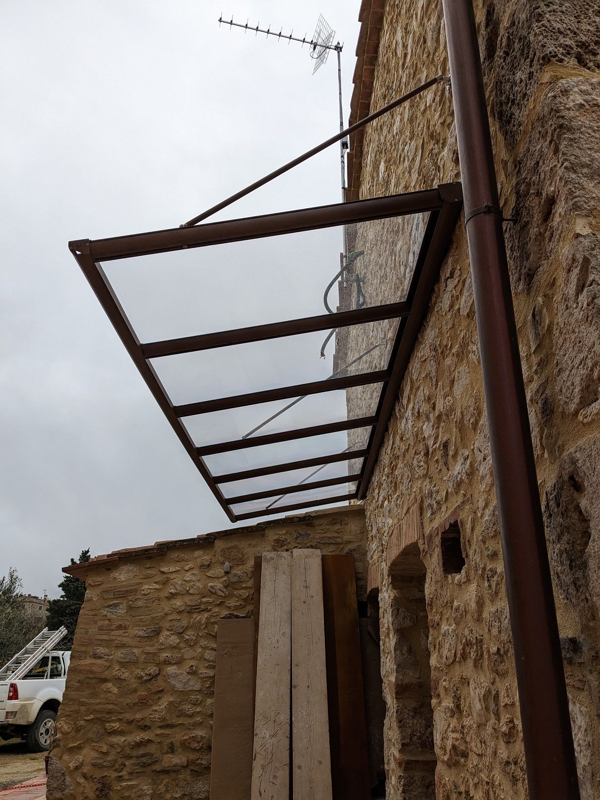 Struttura di tettoia in metallo arrugginito fissata a un edificio in pietra sotto un cielo nuvoloso.