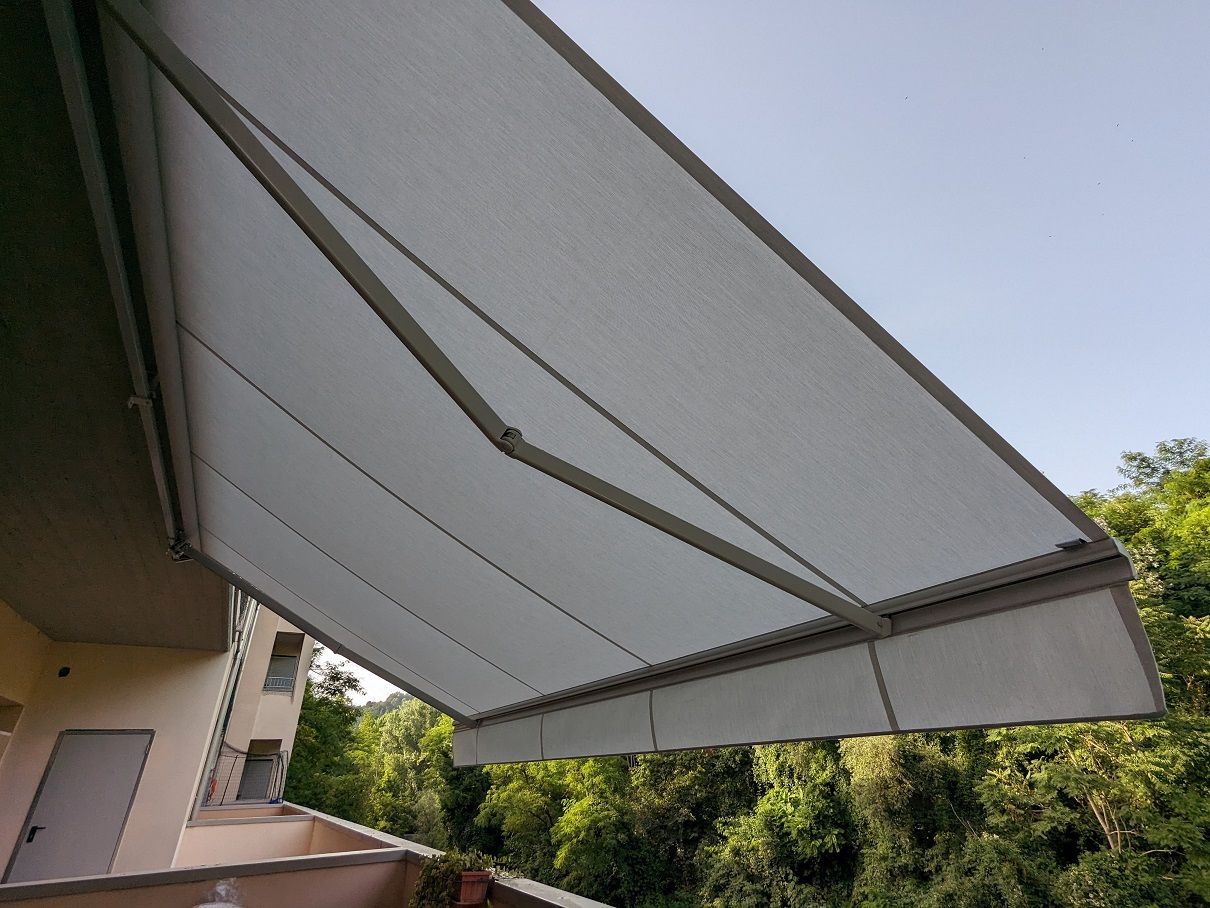 Tenda da sole retrattile grigia che si estende su un patio, con alberi e cielo azzurro sullo sfondo.