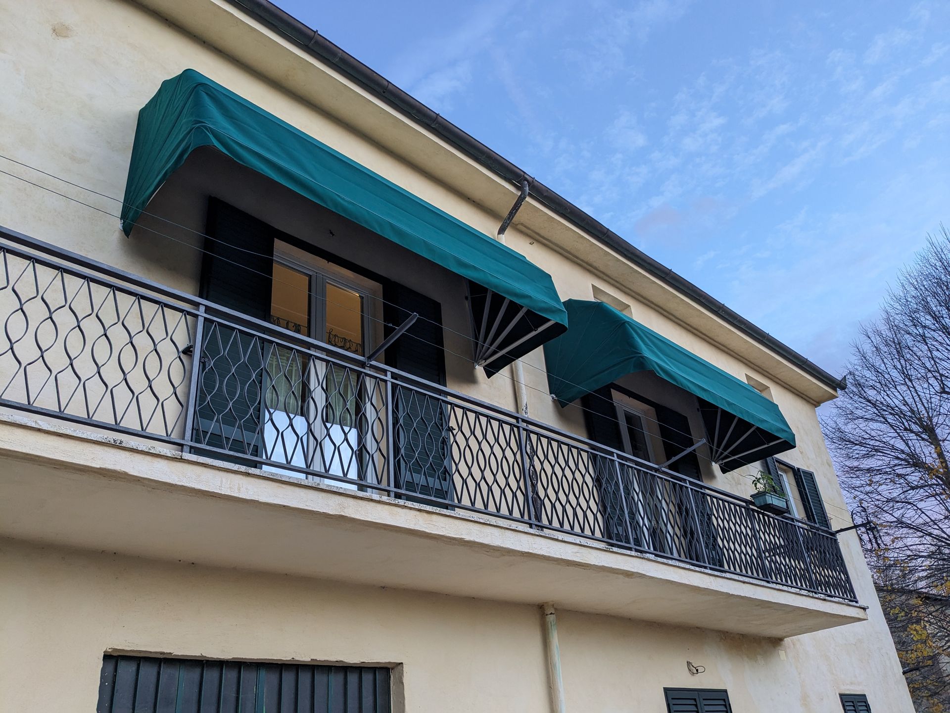 Edificio beige a due piani con balcone in ferro battuto nero e tende da sole color verde acqua contro un cielo azzurro.