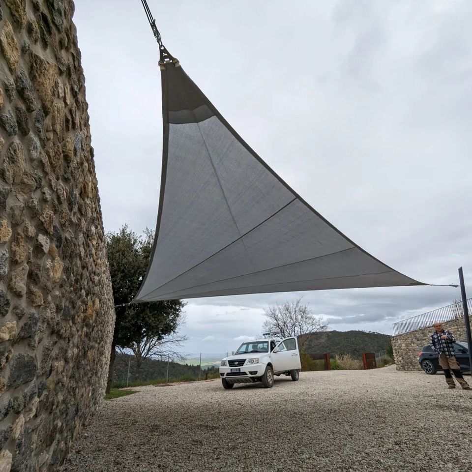Una vela grigia ombreggiante si staglia su un piazzale ghiaioso con un furgone bianco, un muro di pietra e due persone vicino a una tenda.