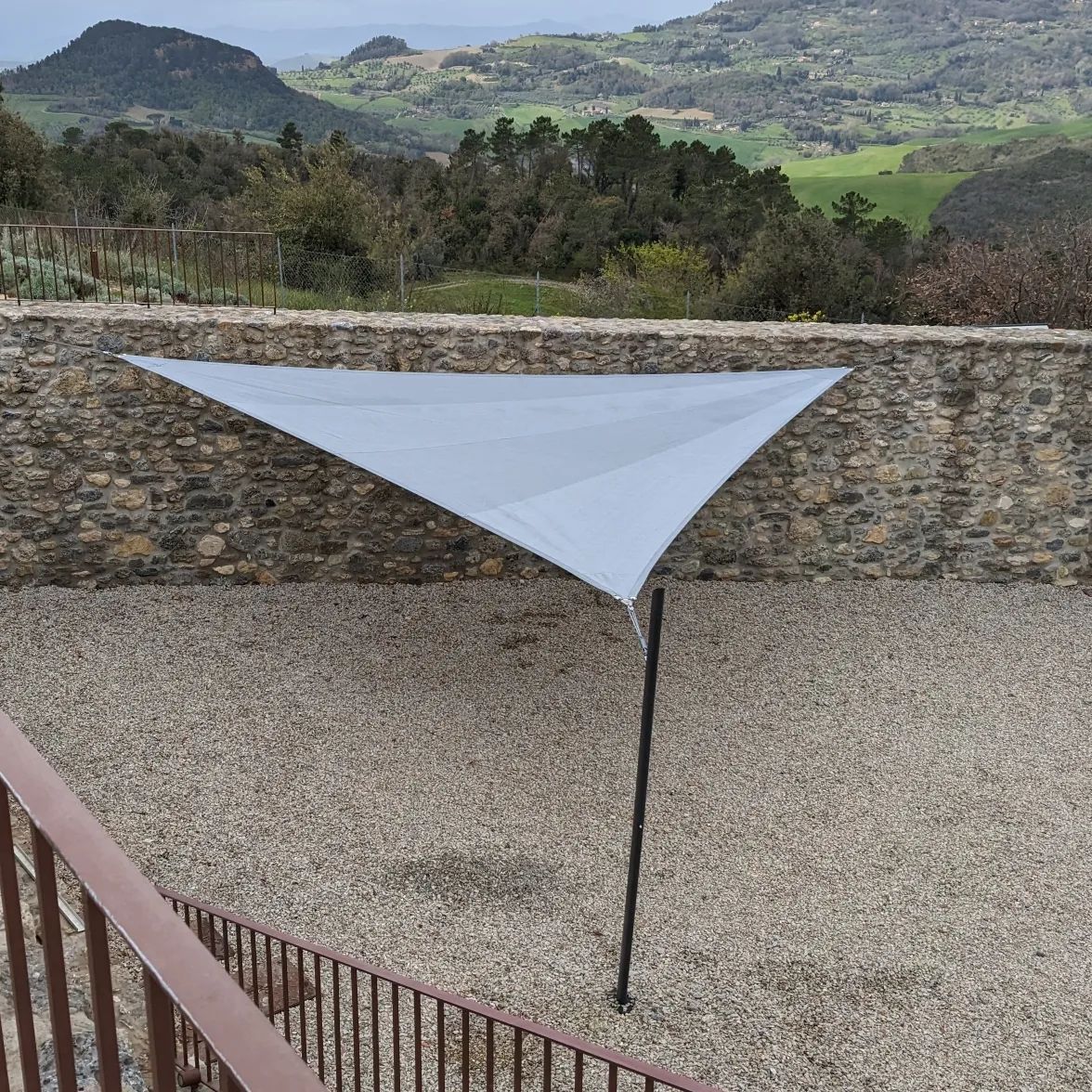 Tenda parasole triangolare bianca su un palo in un cortile di ghiaia, con un muro di pietra e verdi colline sullo sfondo.