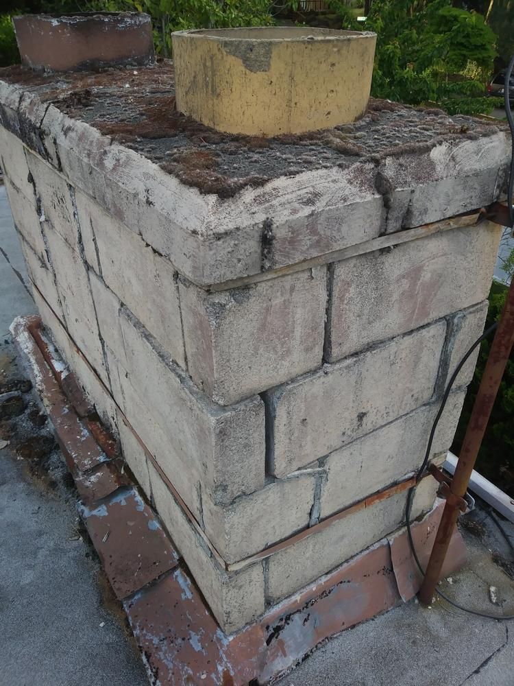 Weathered brick chimney stack on a rooftop, with a yellow cap and surrounding debris.