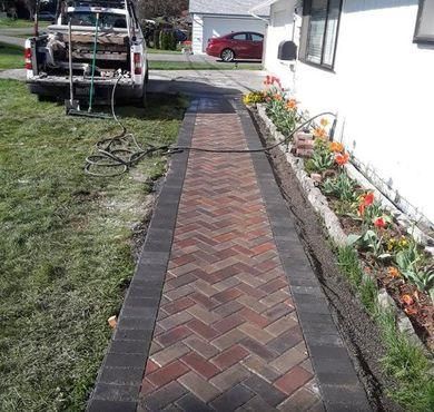 Brick garden path beside a white house, bordered by flowers and grass, leading to a parked truck.