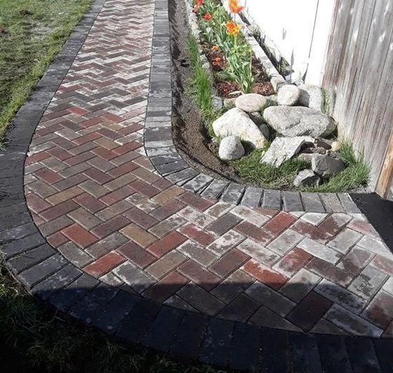 Curved brick garden path bordered by dark edging, with stones and flowers along a white wall