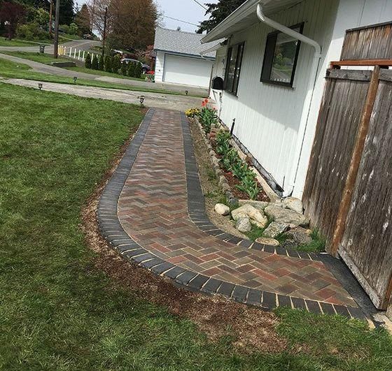 Curved brick walkway beside a white house and wooden fence, leading to a driveway and garage.
