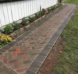 Paved brick walkway beside a white wall and garden bed, bordered by dark edging.